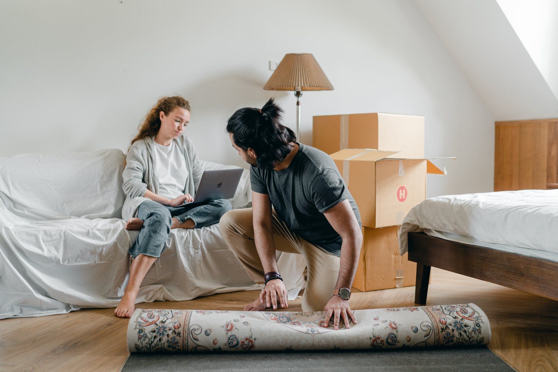 Couple in a bright room: woman using a laptop, man rolling out a rug, boxes and a lamp nearby.