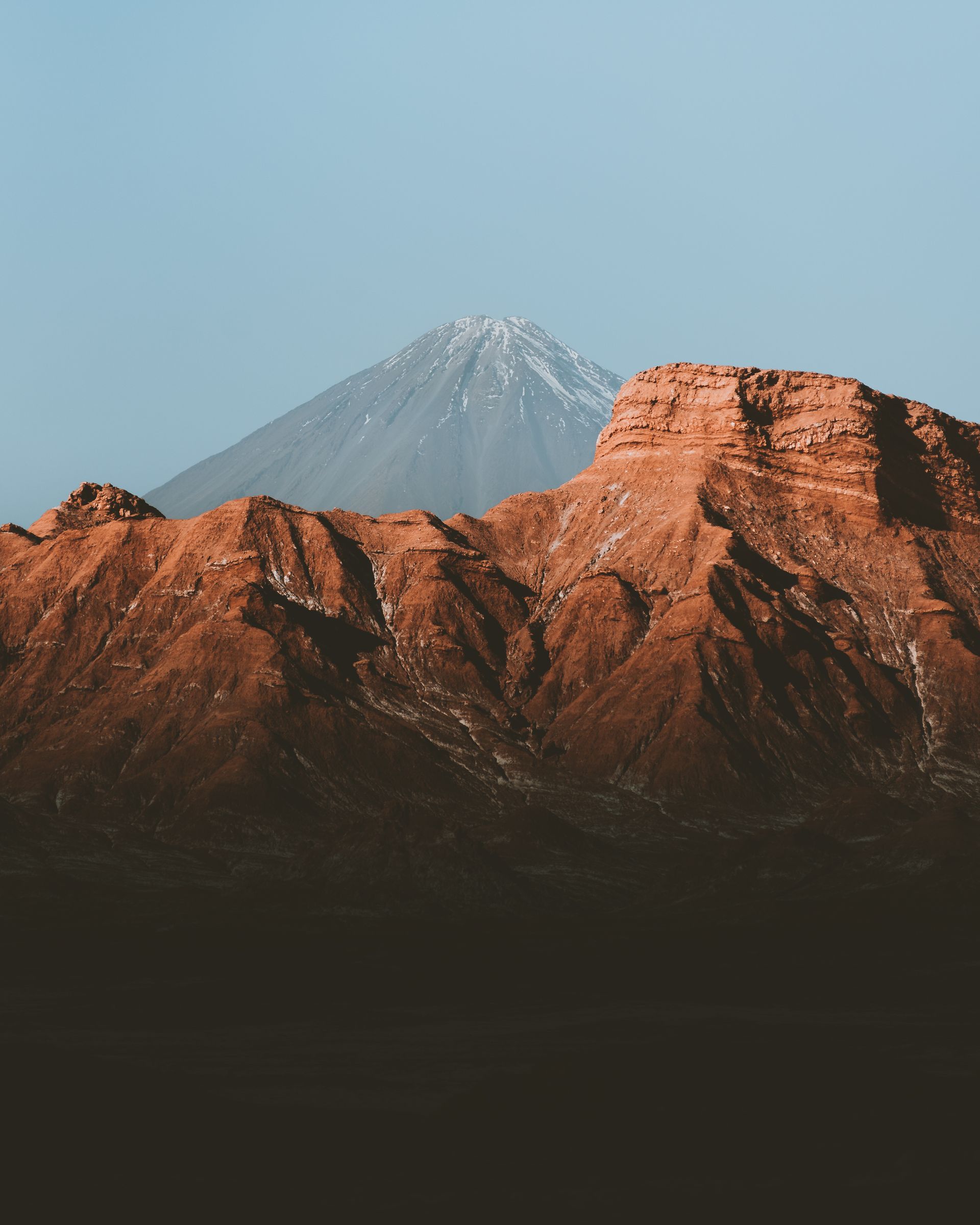 Pico de montaña cubierto de nieve detrás de acantilados desérticos de color marrón rojizo bajo un cielo azul claro.