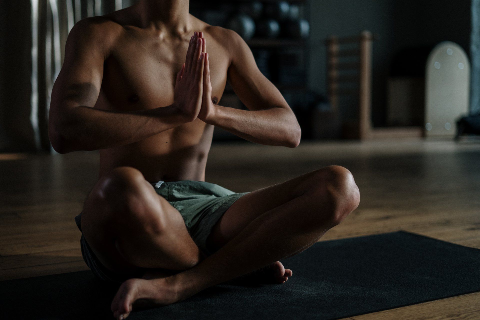 A shirtless man is sitting on a yoga mat with his hands folded in prayer.