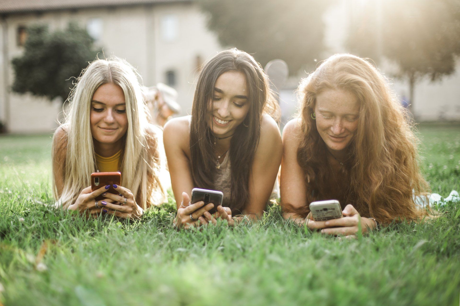 three women are laying in the grass looking at their cell phones .