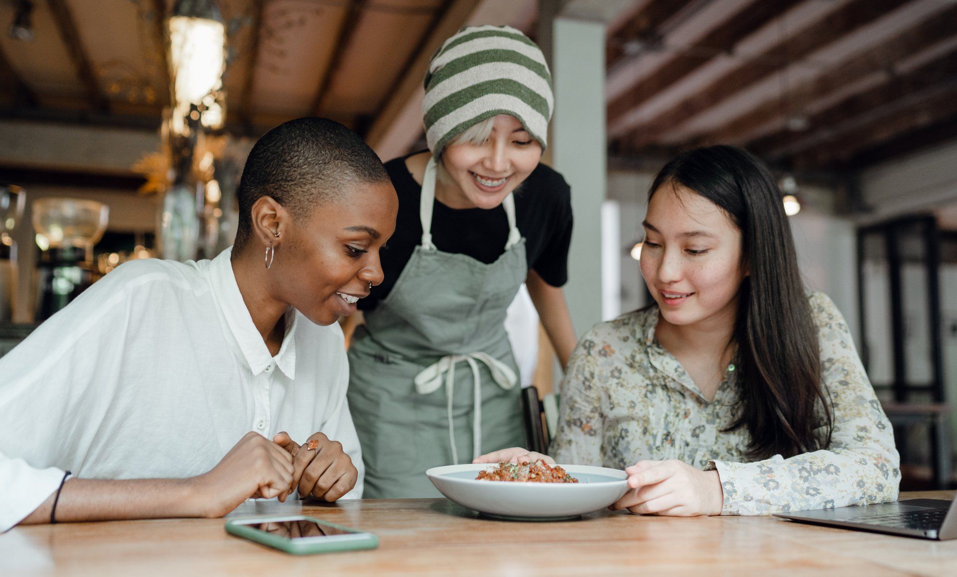 Three people at a table in a cafe; a server presents a dish to two customers.