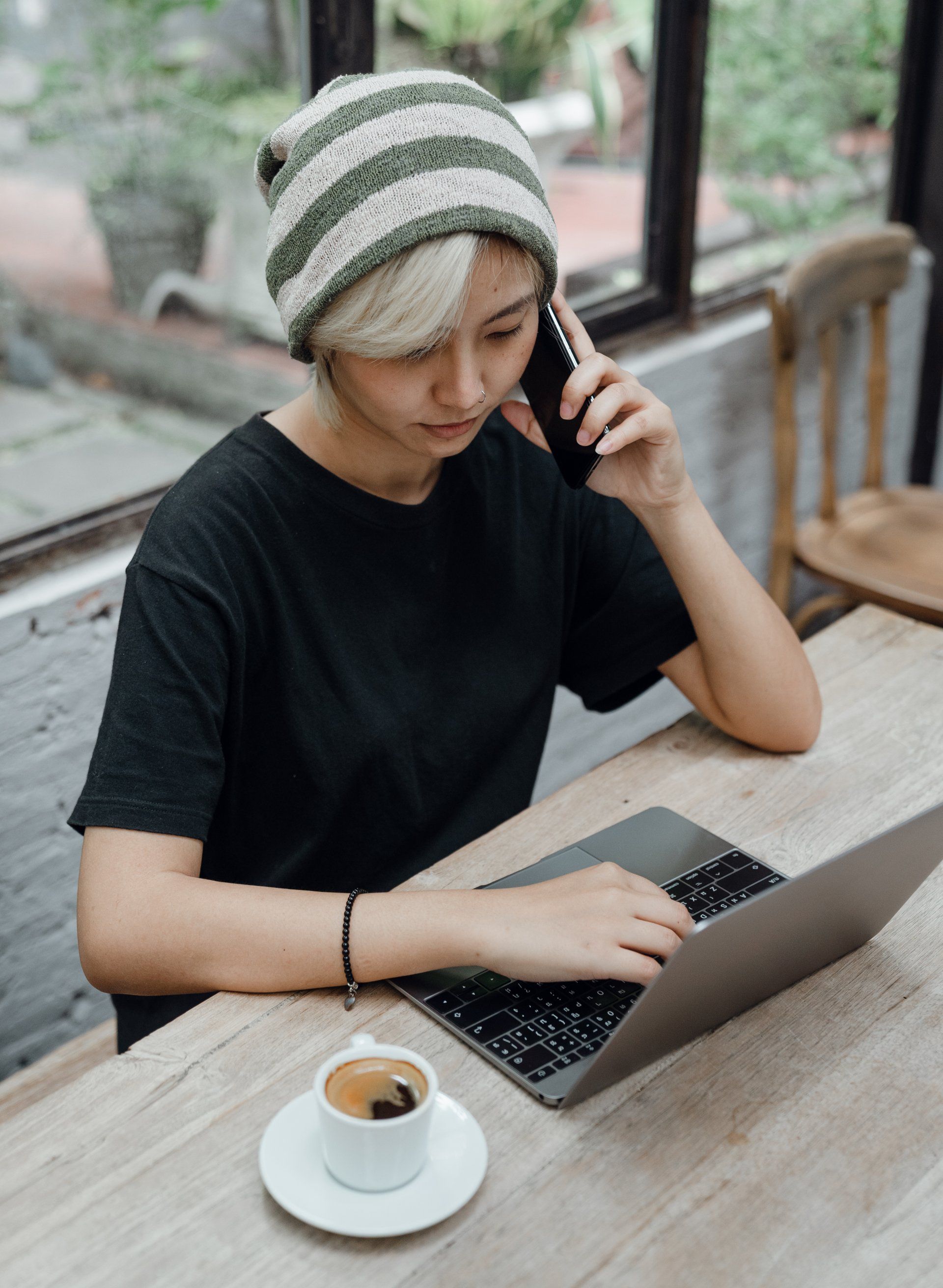 A woman is sitting at a table using a laptop and talking on a cell phone.