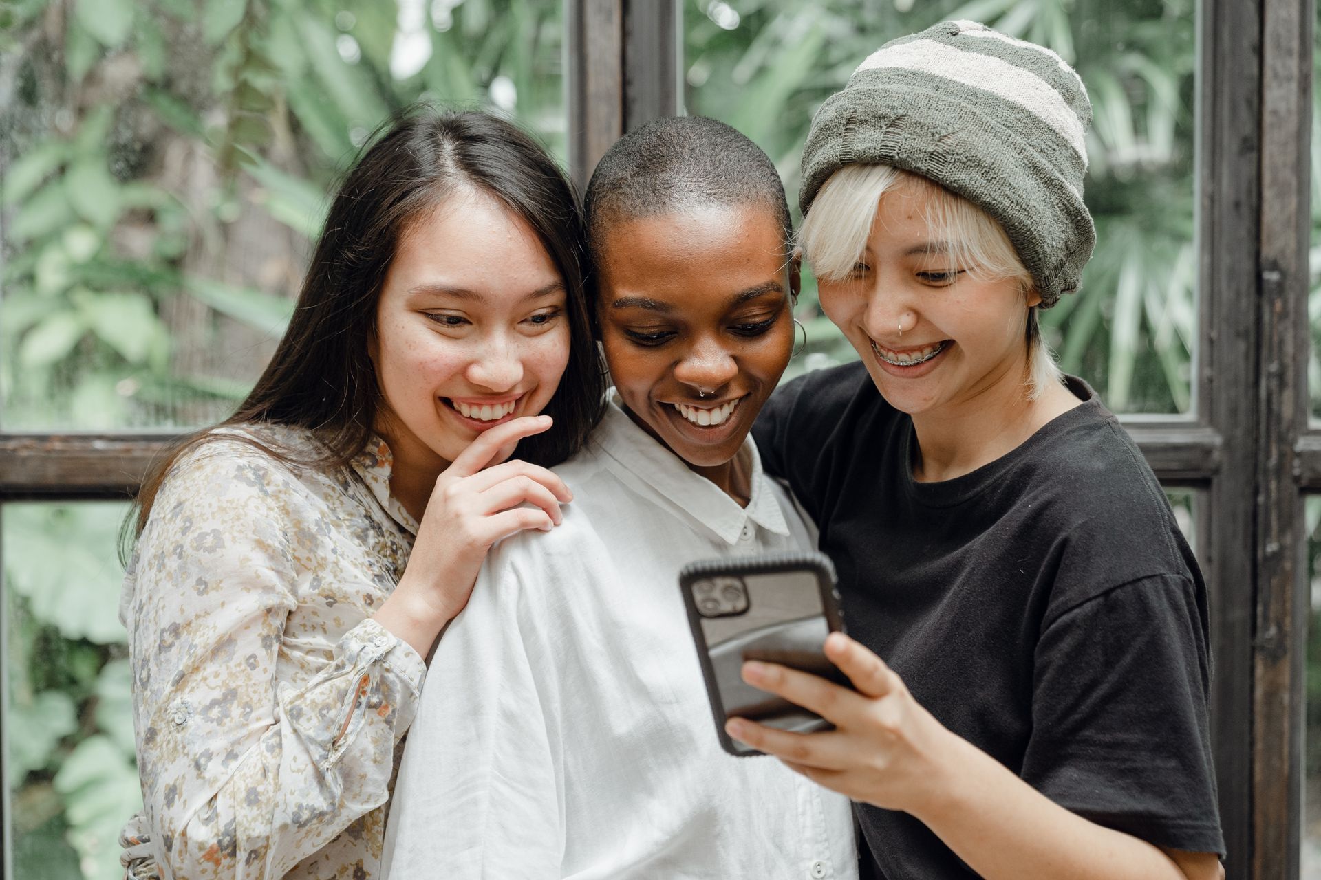 Three diverse women smiling, looking at a phone together by a window.