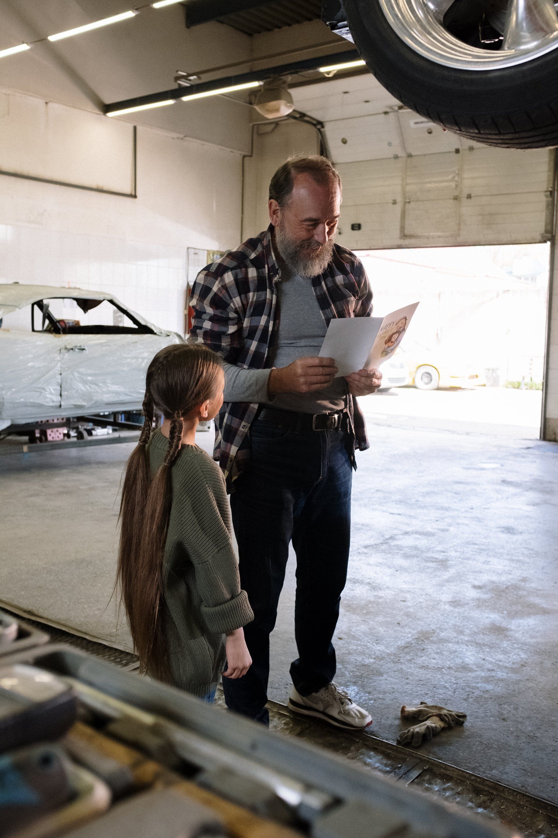 A man and a little girl are standing next to a car in a garage.