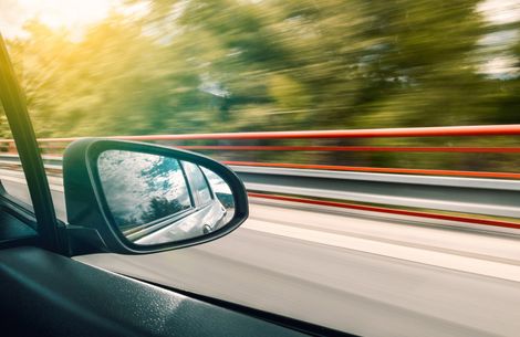 Car side mirror showing blurred scenery of road and trees, reflecting blue sky.
