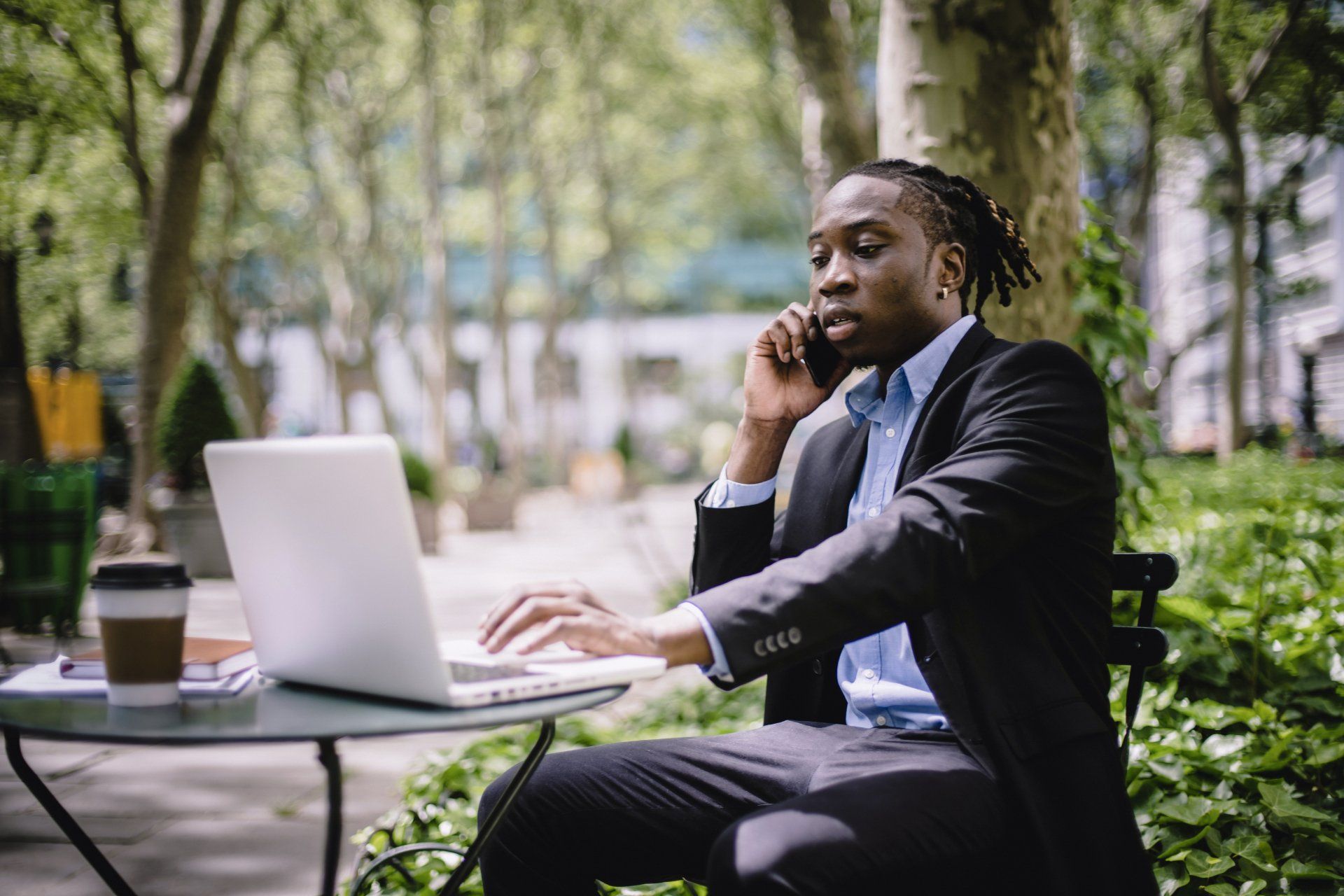 A man in a suit is sitting at a table with a laptop and talking on a cell phone.