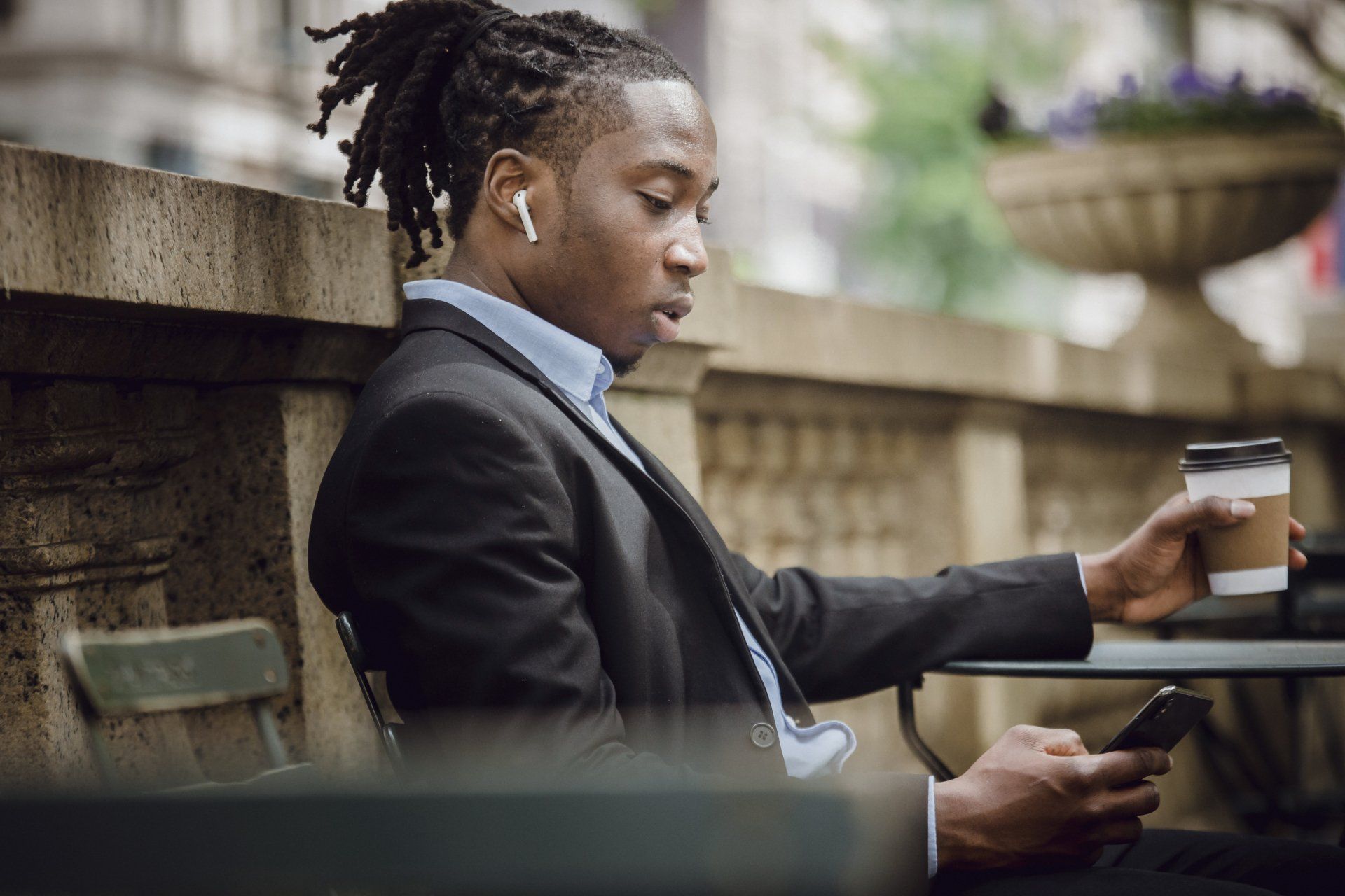 A man in a suit is sitting at a table holding a cup of coffee and looking at his phone.