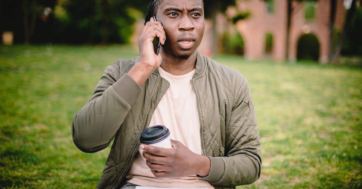 A man is sitting on the grass talking on a cell phone while holding a cup of coffee.