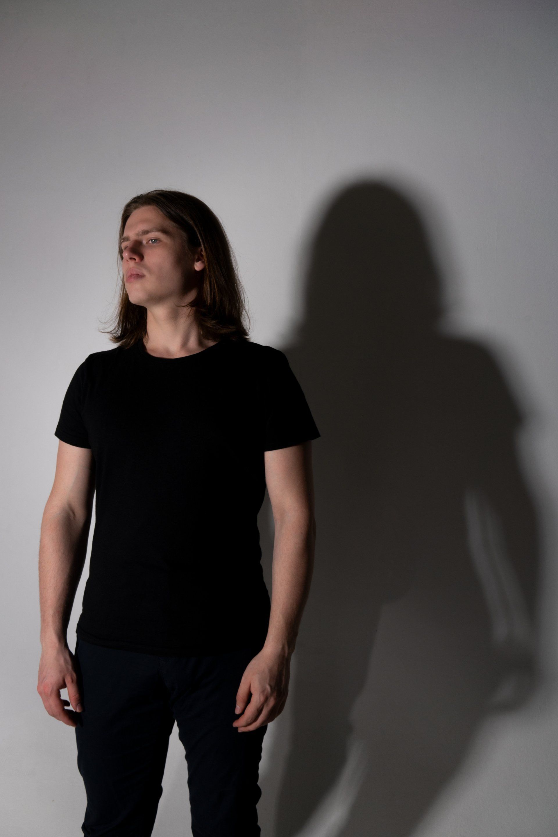 A man in a black shirt is standing in front of a white wall with his shadow on the wall.