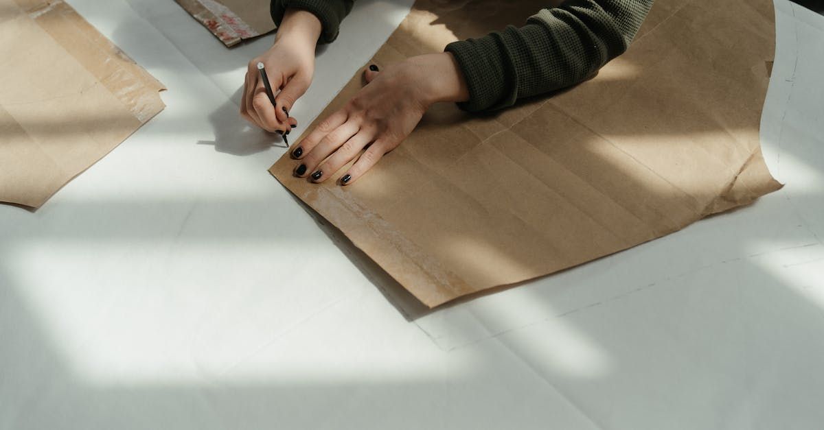 De handen van een persoon tekenen op bruin papier dat op een wit oppervlak ligt, belicht door zonlicht.
