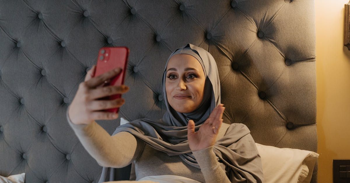 A woman in a hijab is taking a selfie with her phone while sitting on a bed.