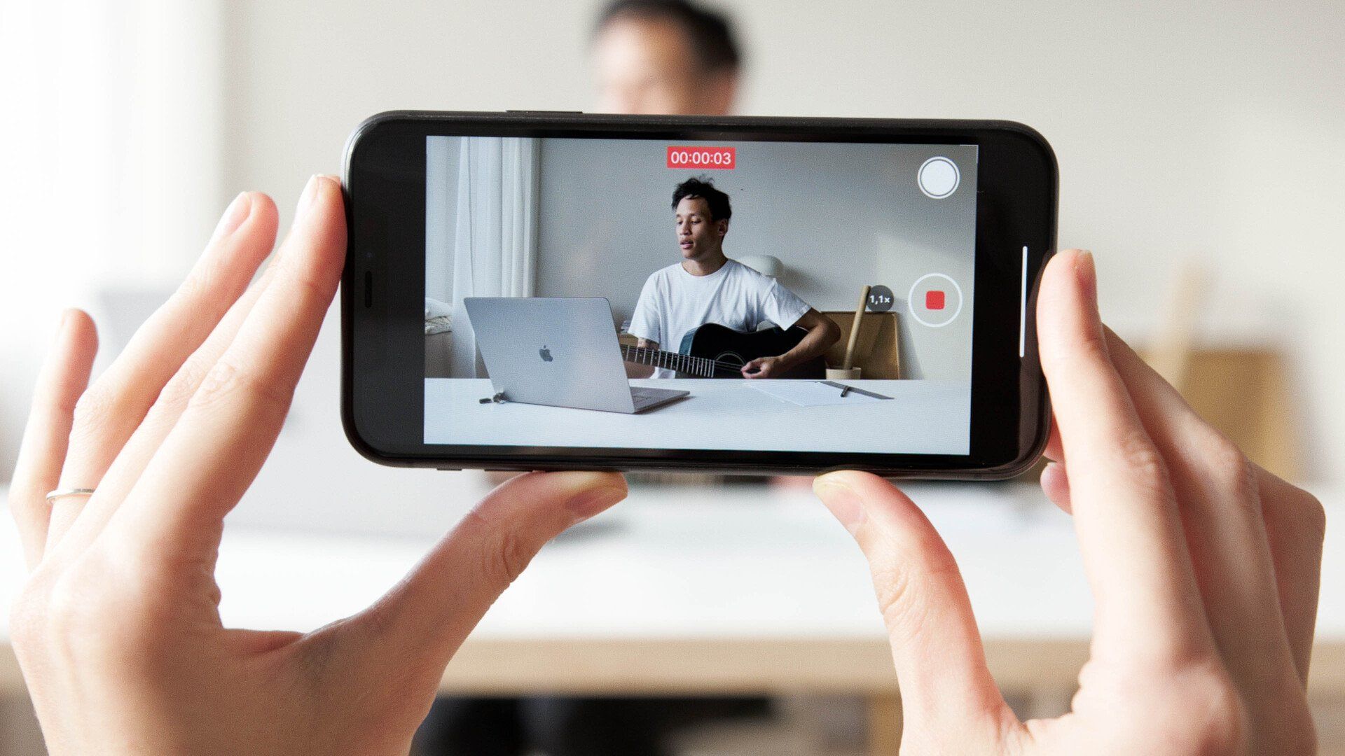 A person is taking a picture of a man sitting at a desk with a laptop.