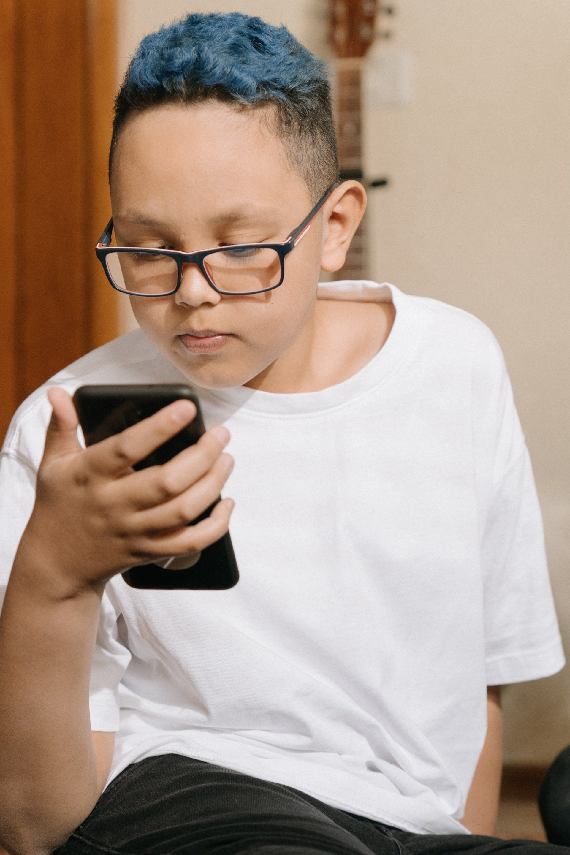 a young boy with blue hair and glasses is using a cell phone .