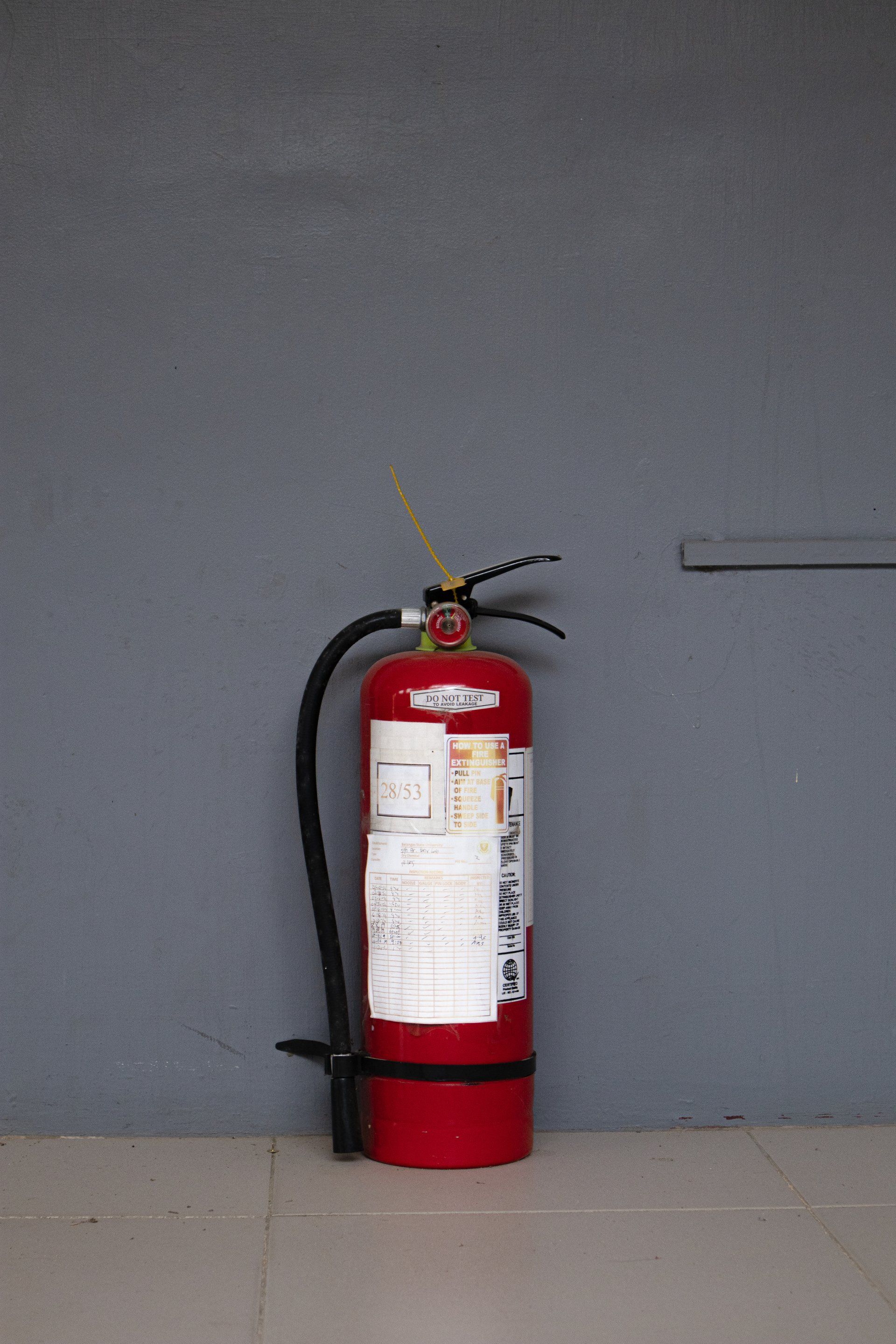 A red fire extinguisher is sitting in front of a gray wall.