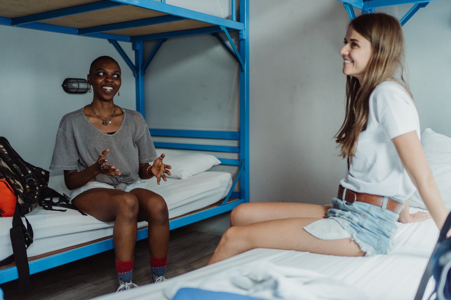 Two young women talking on beds in a hostel room with blue bunk beds. One is black with short hair and the other is white with long hair.