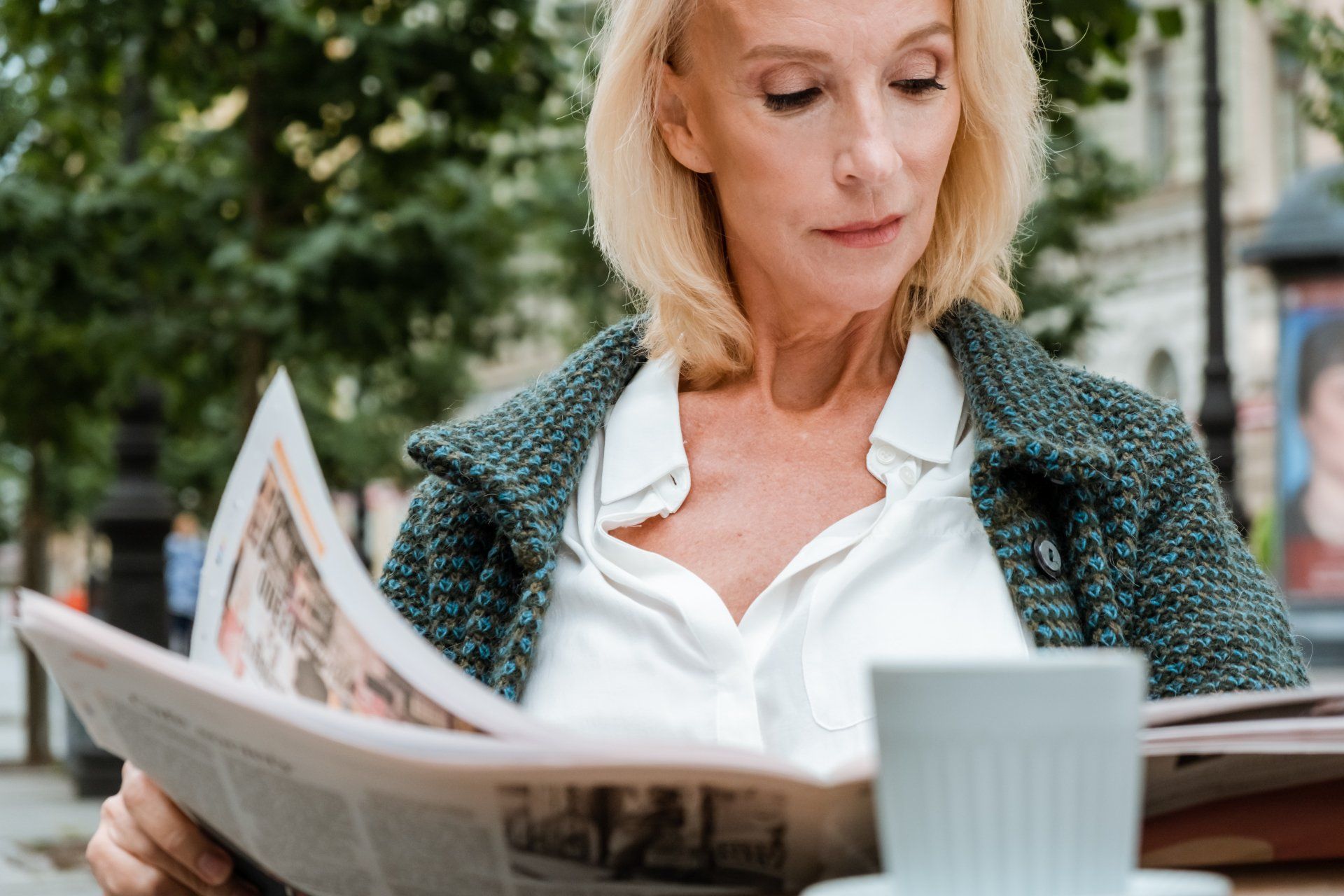 A woman is reading a newspaper while sitting at a table with a cup of coffee.