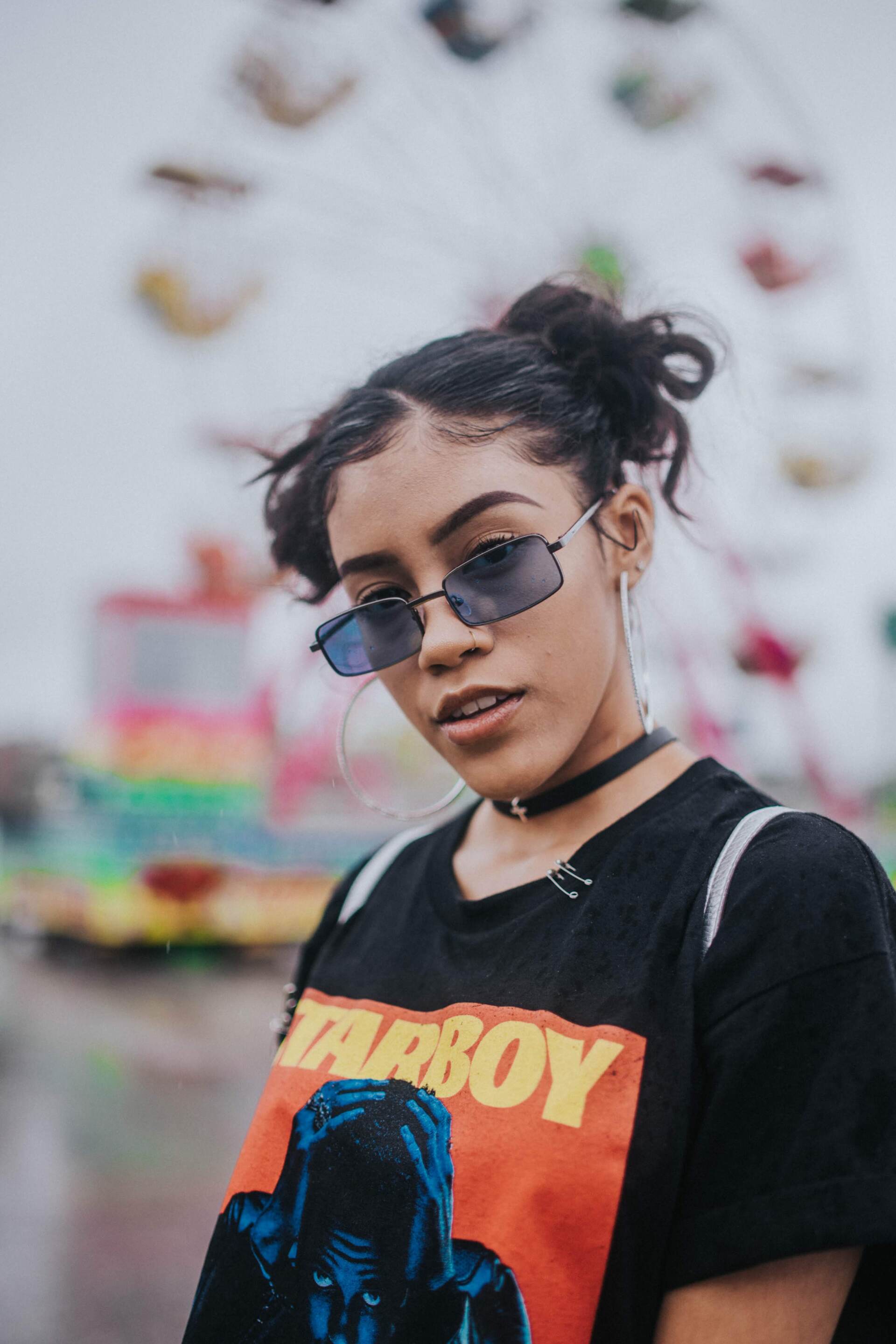 A woman wearing sunglasses and a t-shirt is standing in front of a ferris wheel.
