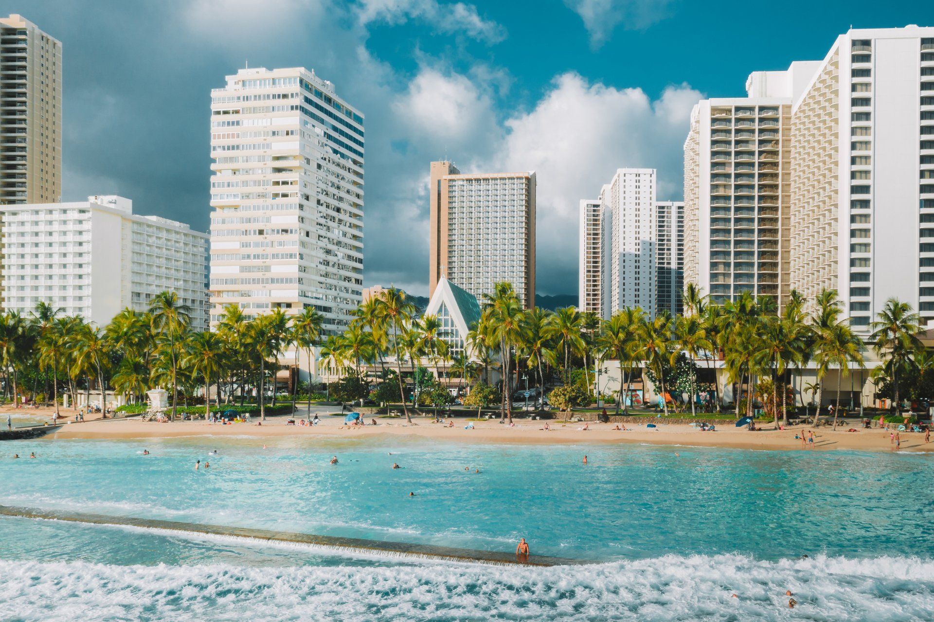 Tall white hotels rise above lush green palm trees along the sandy shore of Waikiki Beach under a cloudy blue sky.