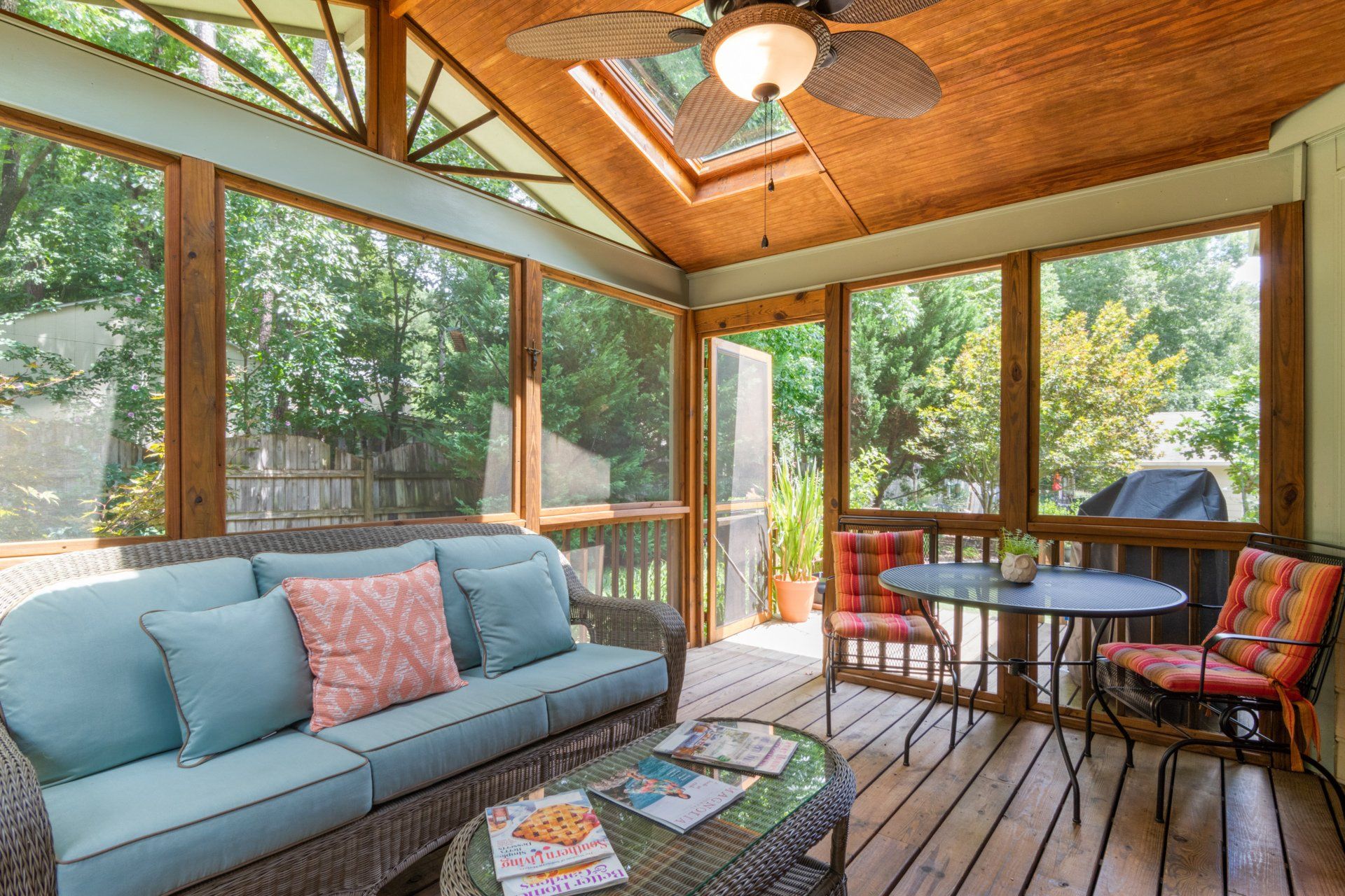 A screened in porch with a couch , table , chairs and a ceiling fan.