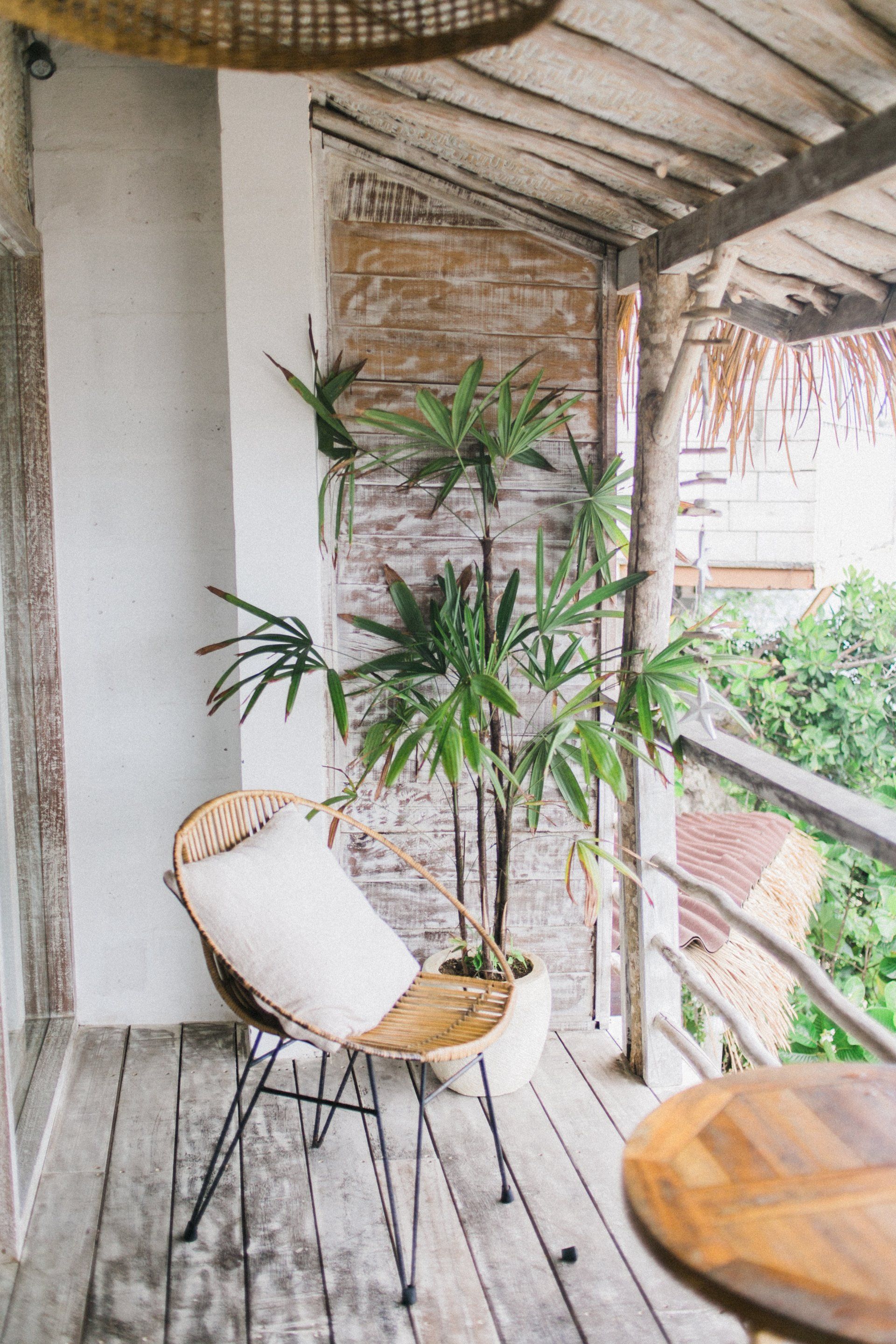 Wicker chair with white cushion on a wooden balcony, tall plant, rustic setting, partial view of surrounding greenery.
