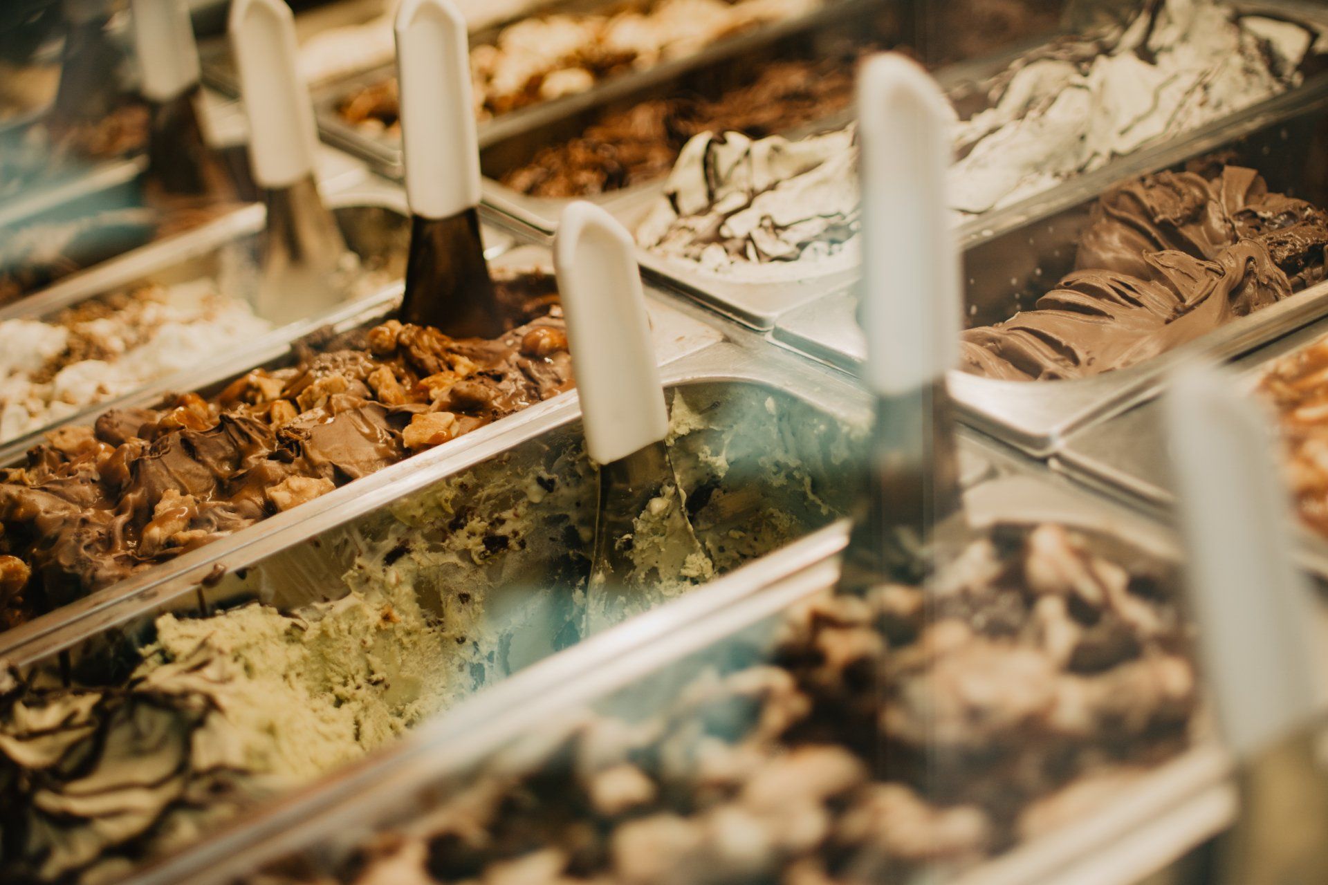 A display case filled with various flavors of gelato in metal containers, each with a serving scoop.
