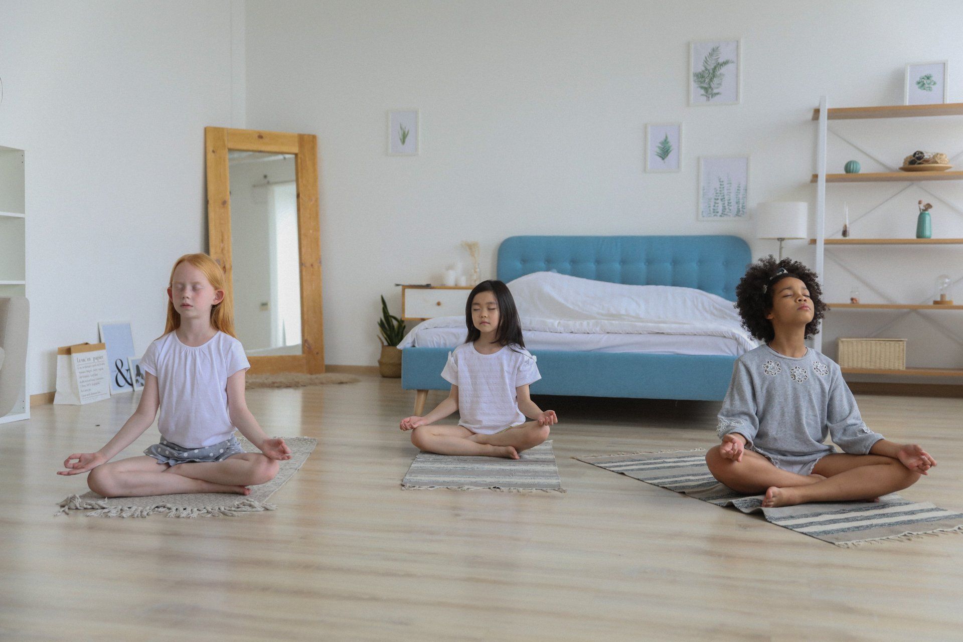 Three children are sitting in a lotus position on yoga mats in a bedroom.