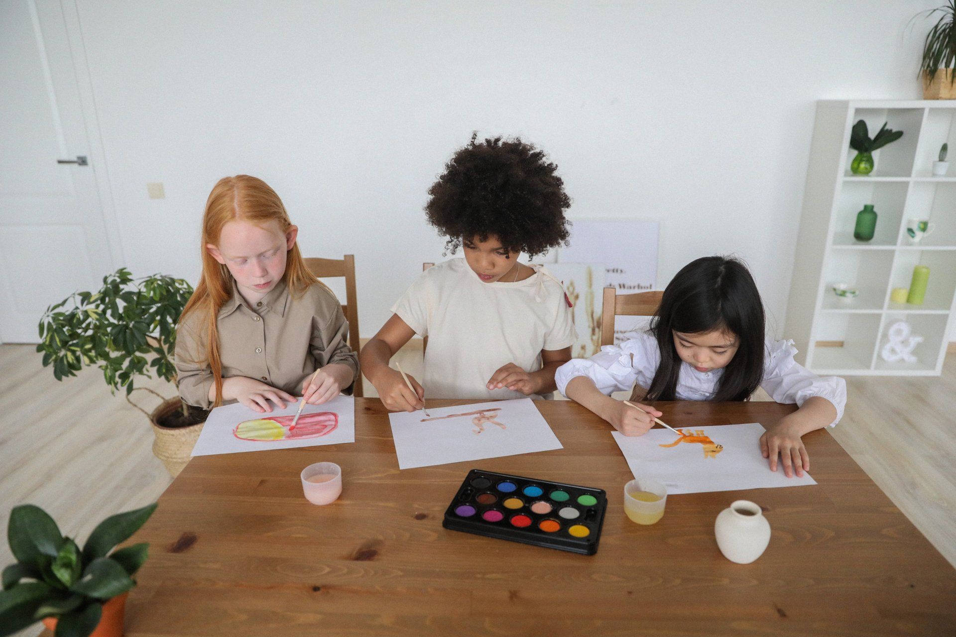 Three children are sitting at a table painting with watercolors.