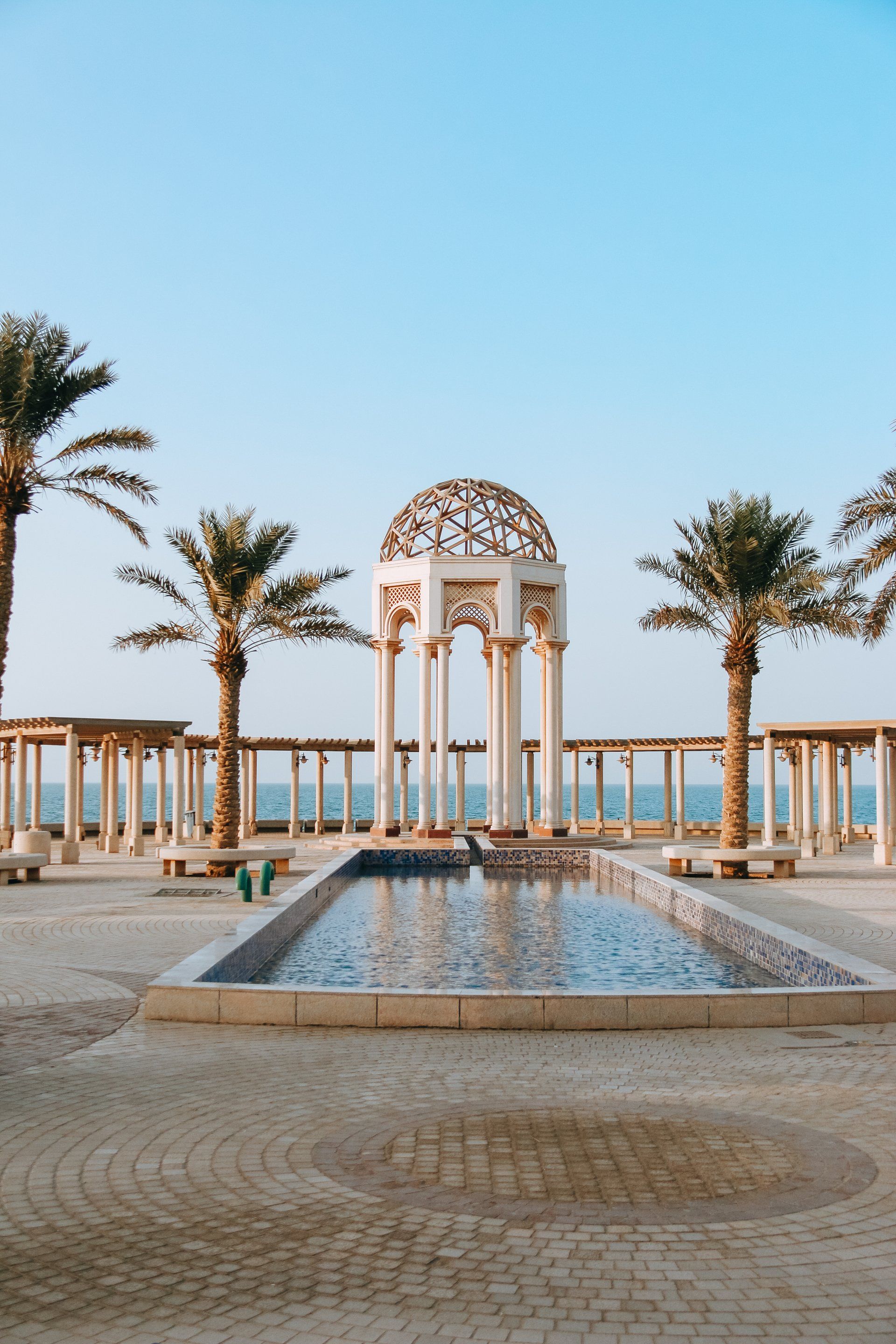 A fountain in a park with palm trees and a gazebo in the background.