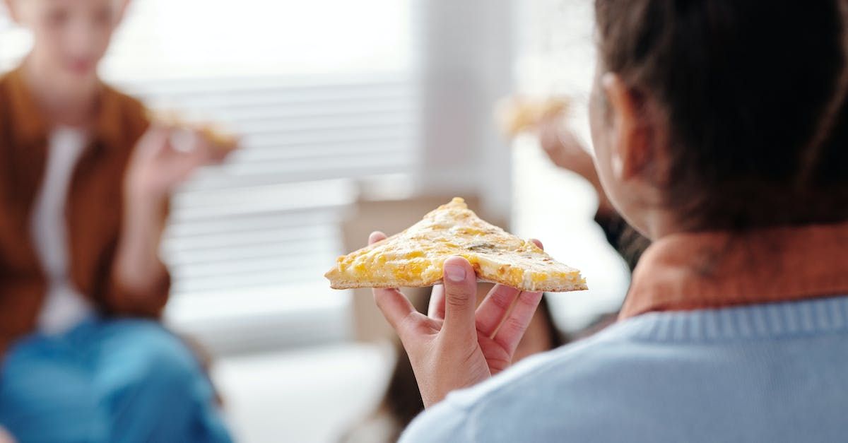 Person holding up a slice of pizza, focus on the pizza. Two other people are blurred in the background, also holding pizza.