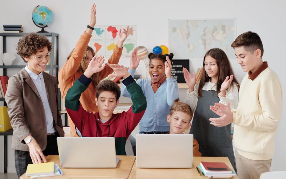 A group of children are standing around two laptops in a classroom.