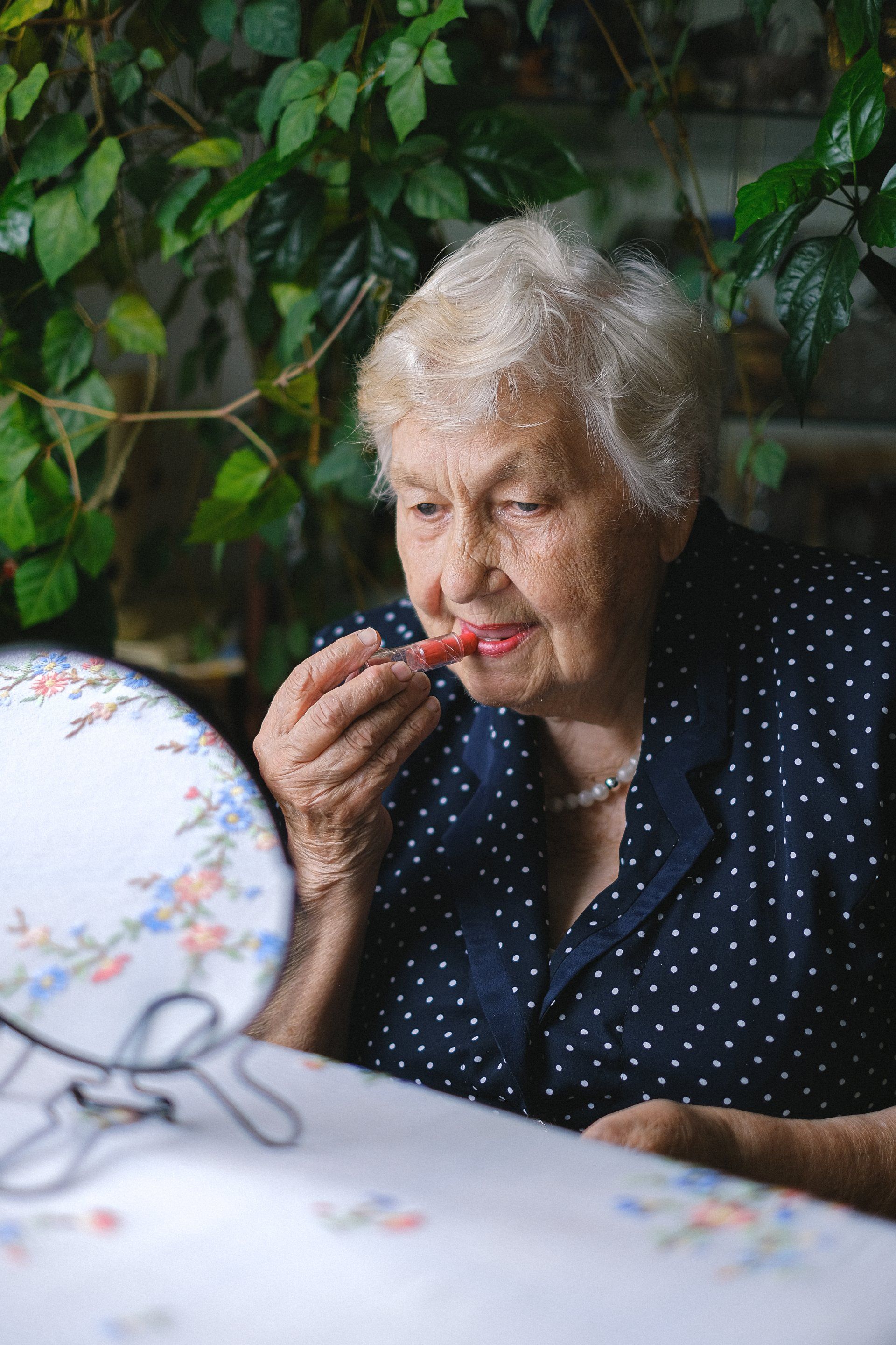 An elderly woman is applying lipstick in front of a mirror.