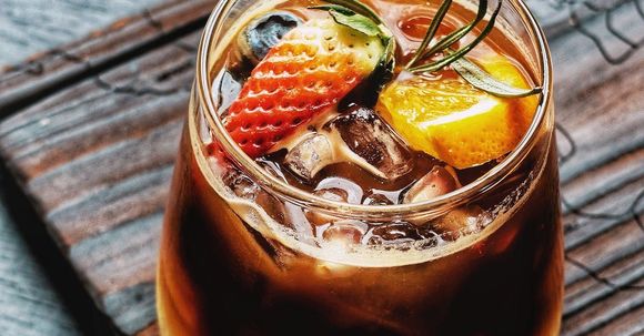 A glass of iced coffee with strawberries , blueberries , orange slices and rosemary on a wooden table.