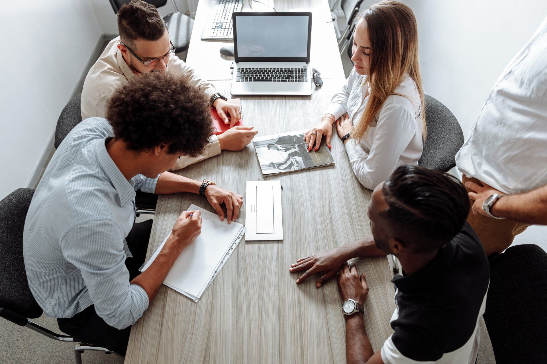 Group of people in a meeting around a table, laptop in the center, discussing ideas, and taking notes.