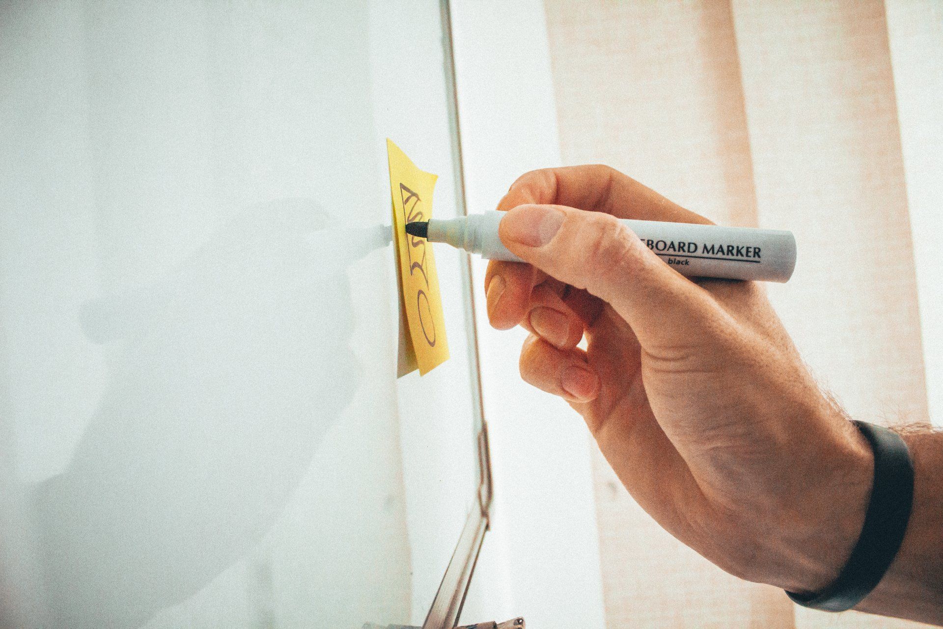 A person draws with a marker on a yellow sticky note attached to a whiteboard.
