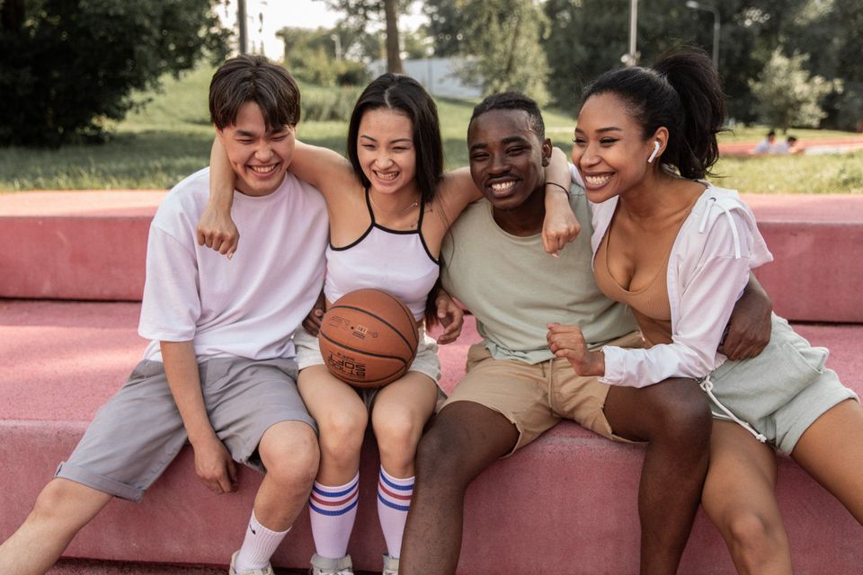 Four diverse teens smiling and embracing outdoors with a basketball.