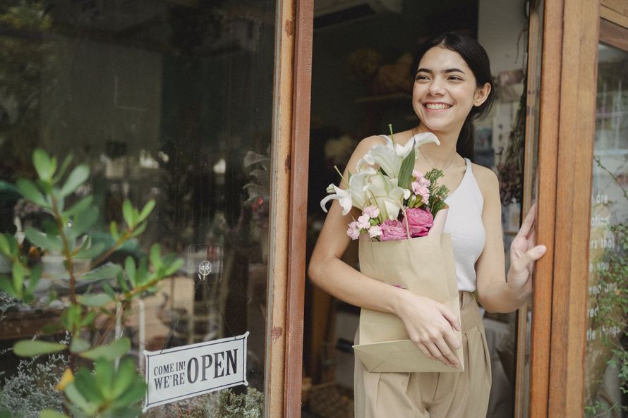 A woman is holding a bag of flowers while standing in front of a flower shop.