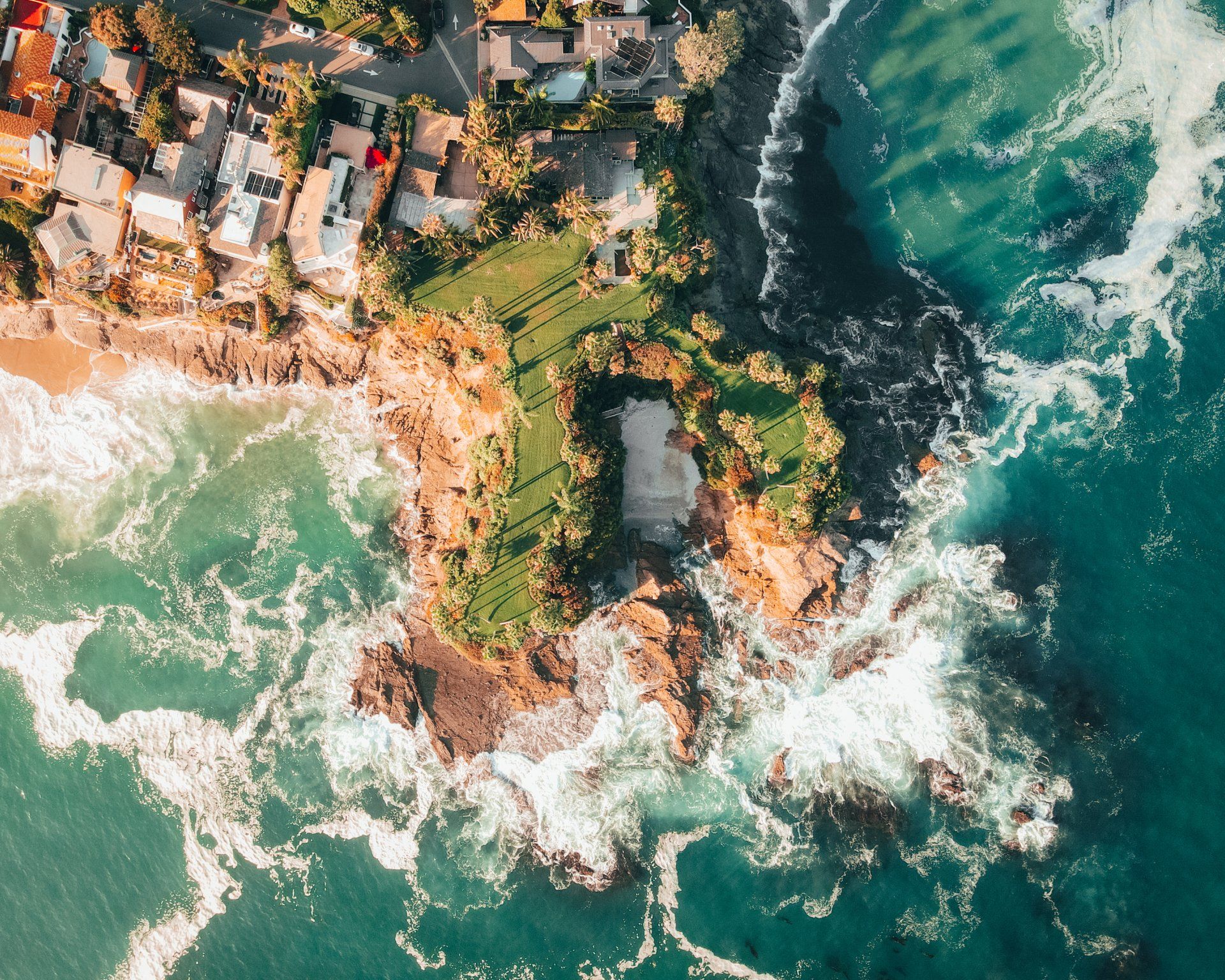 Aerial view of coastal homes and a grassy area surrounded by turquoise ocean with white waves crashing on rocks.