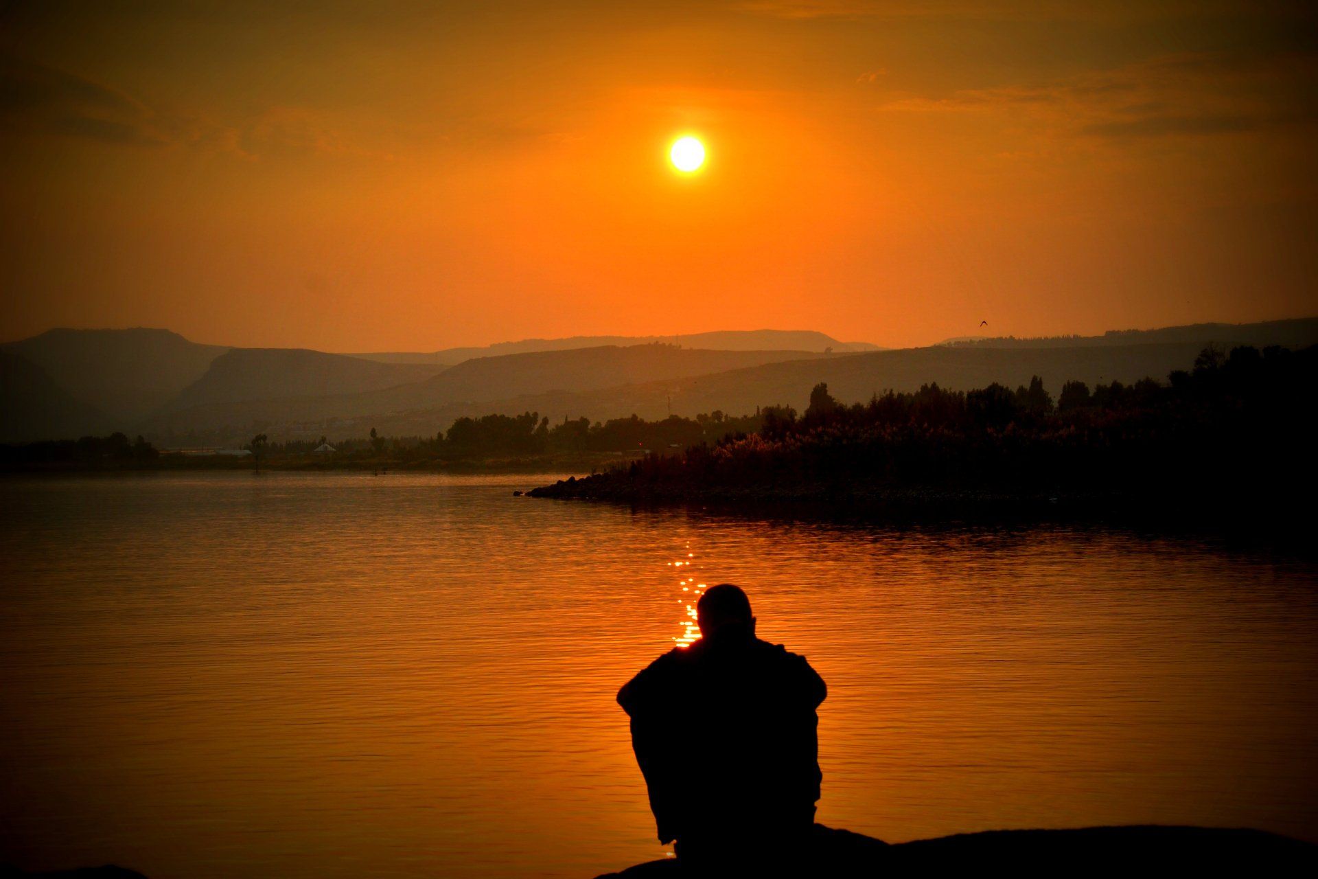 A person is sitting on the shore of a lake at sunset.