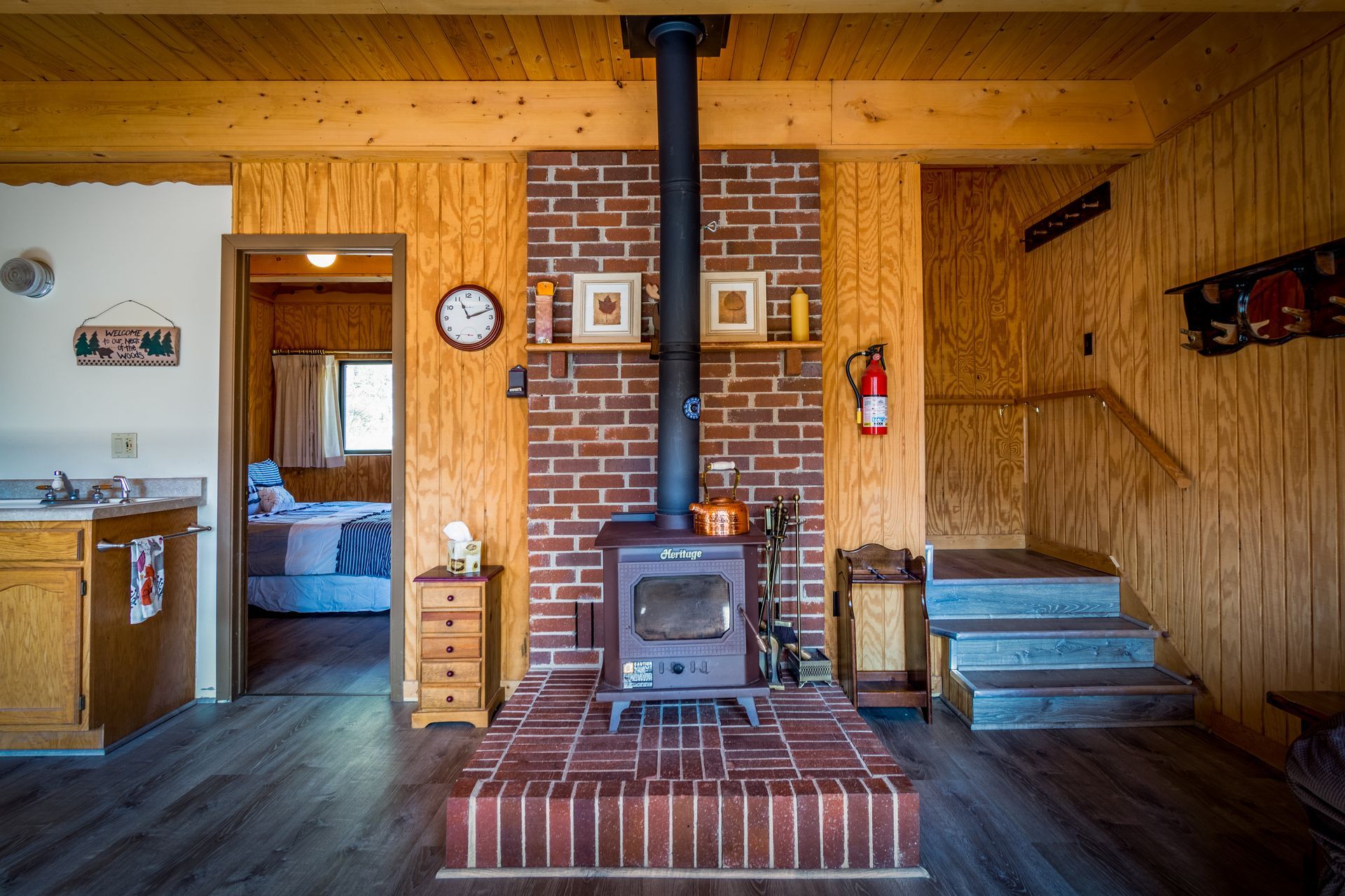 Cozy cabin interior: wood stove on brick, wood-paneled walls, stairs, and a doorway to a bedroom.