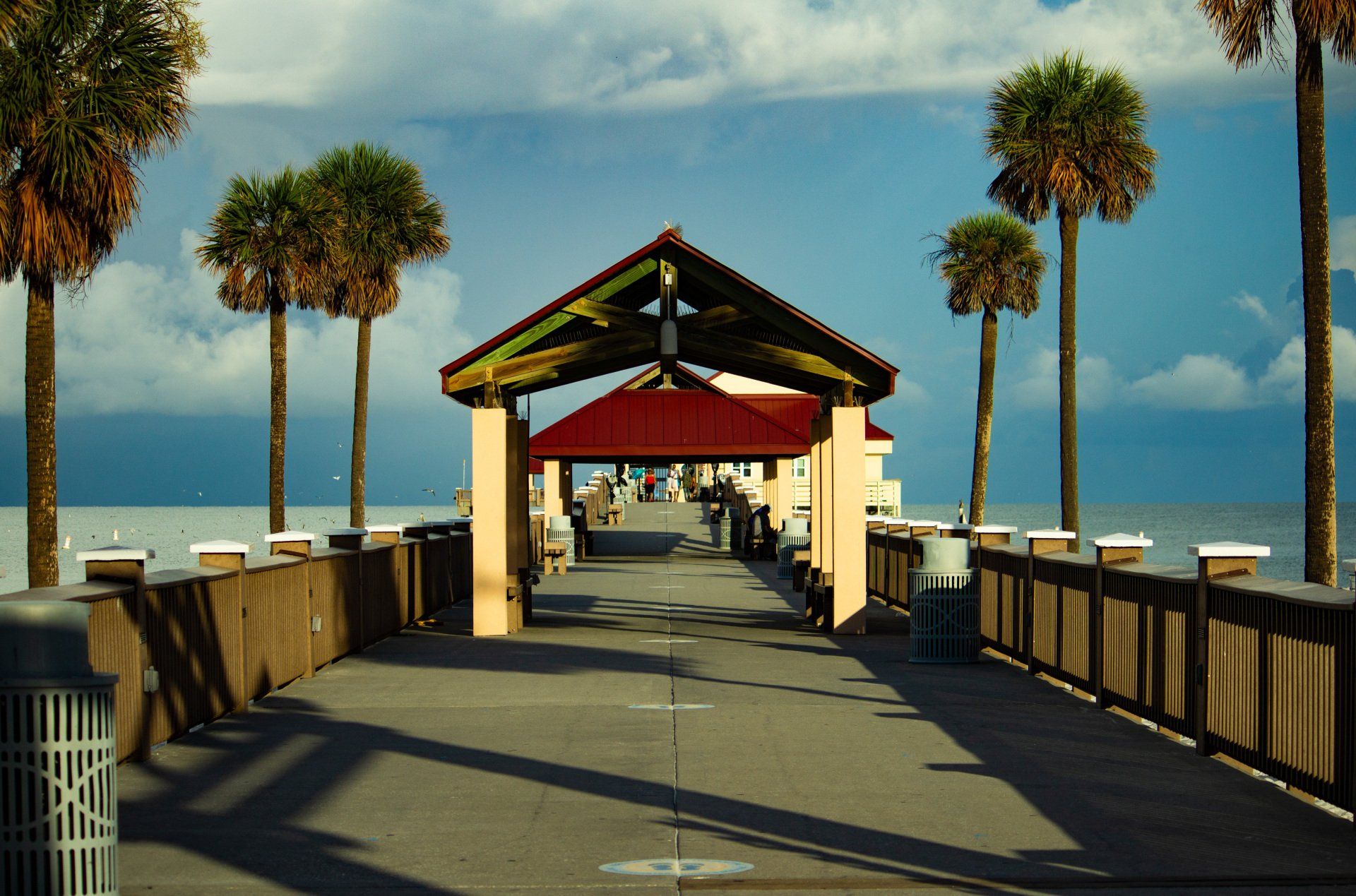 Pier extending into the ocean, lined with palm trees and railings. Red roofed structure.