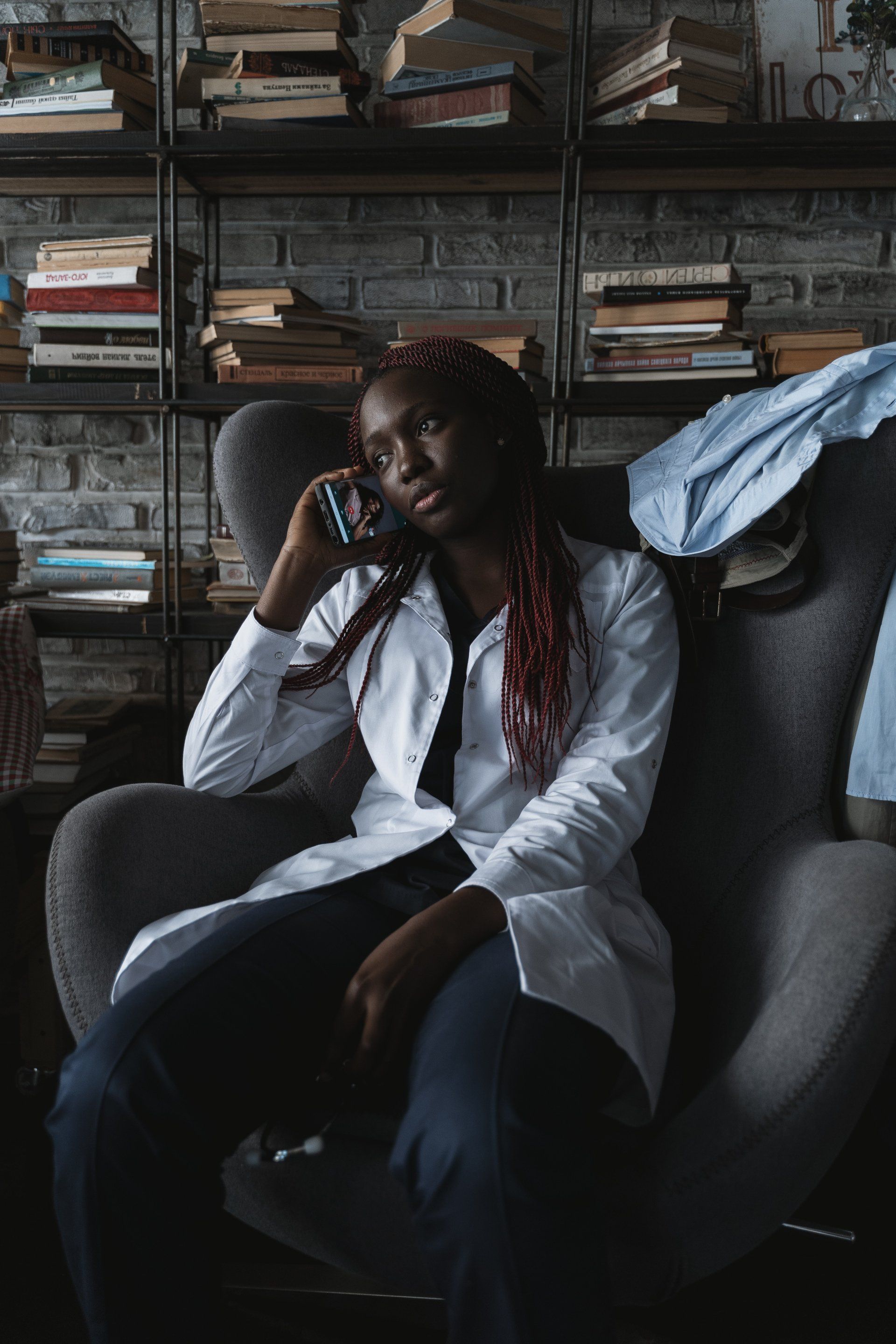 Woman with red-streaked hair in a white coat, talking on a phone while seated in a gray armchair near a bookshelf.