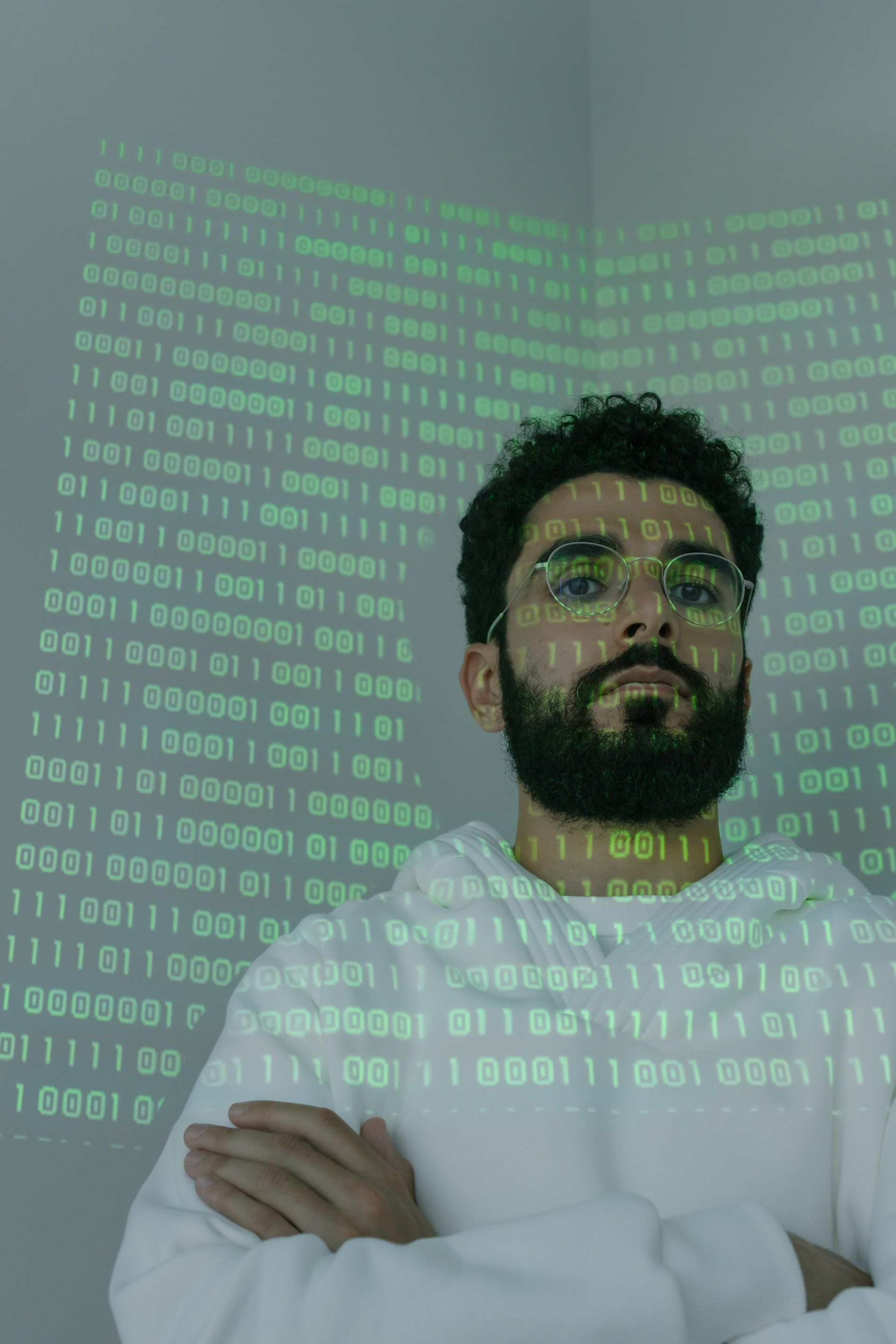 A man with a beard and glasses is standing in front of a wall of binary code.