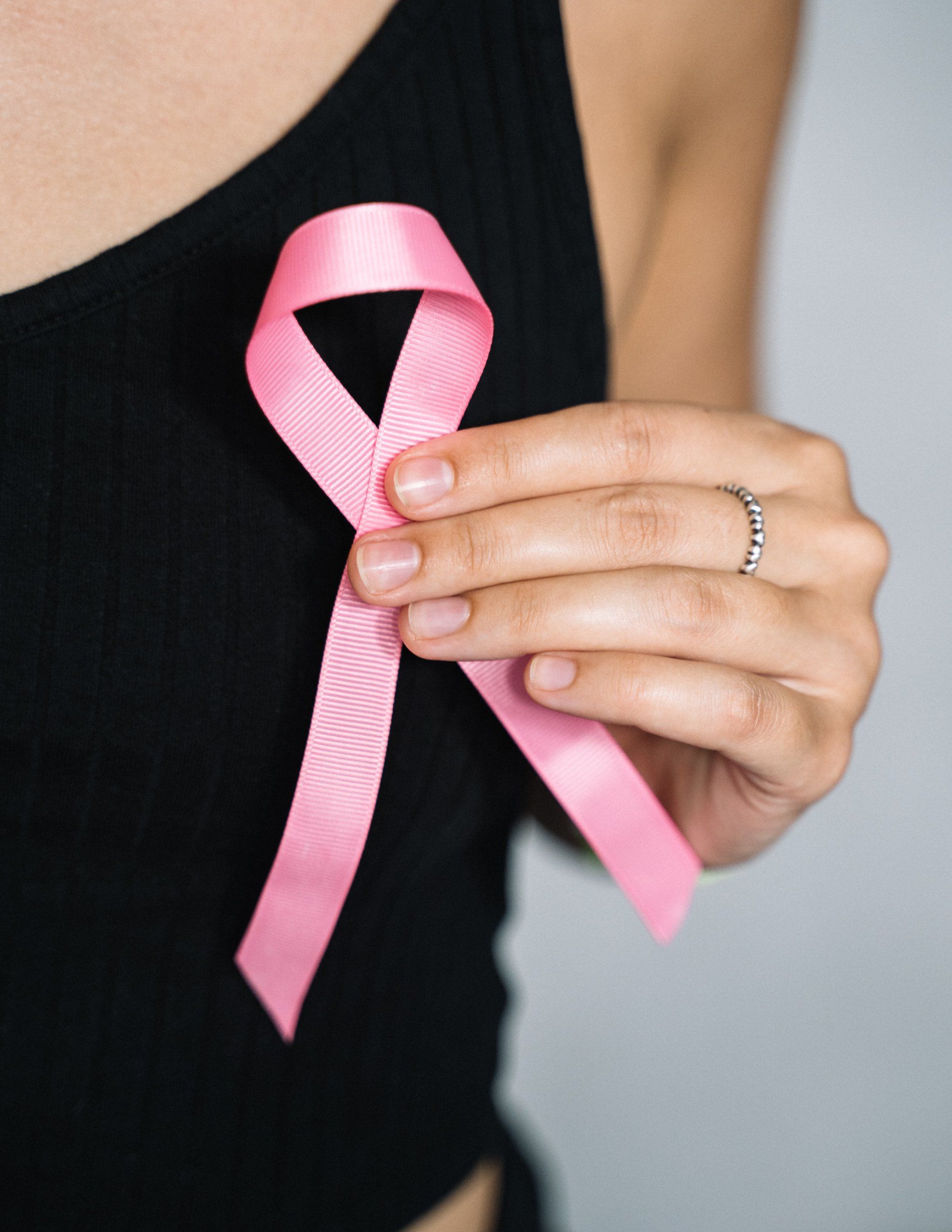 Woman wearing a black top, holding a pink ribbon on her chest, symbolizing breast cancer awareness.