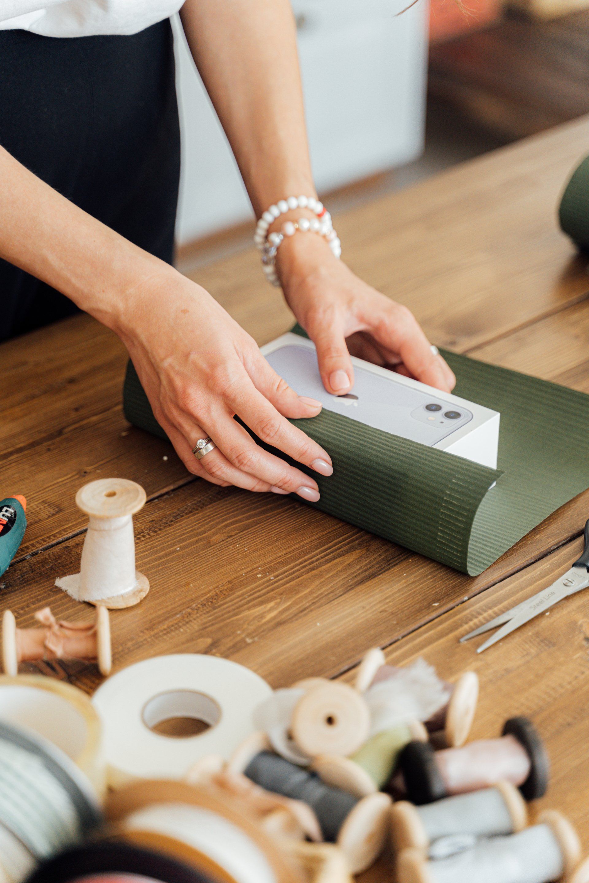 A woman is wrapping a cell phone in green paper on a wooden table.