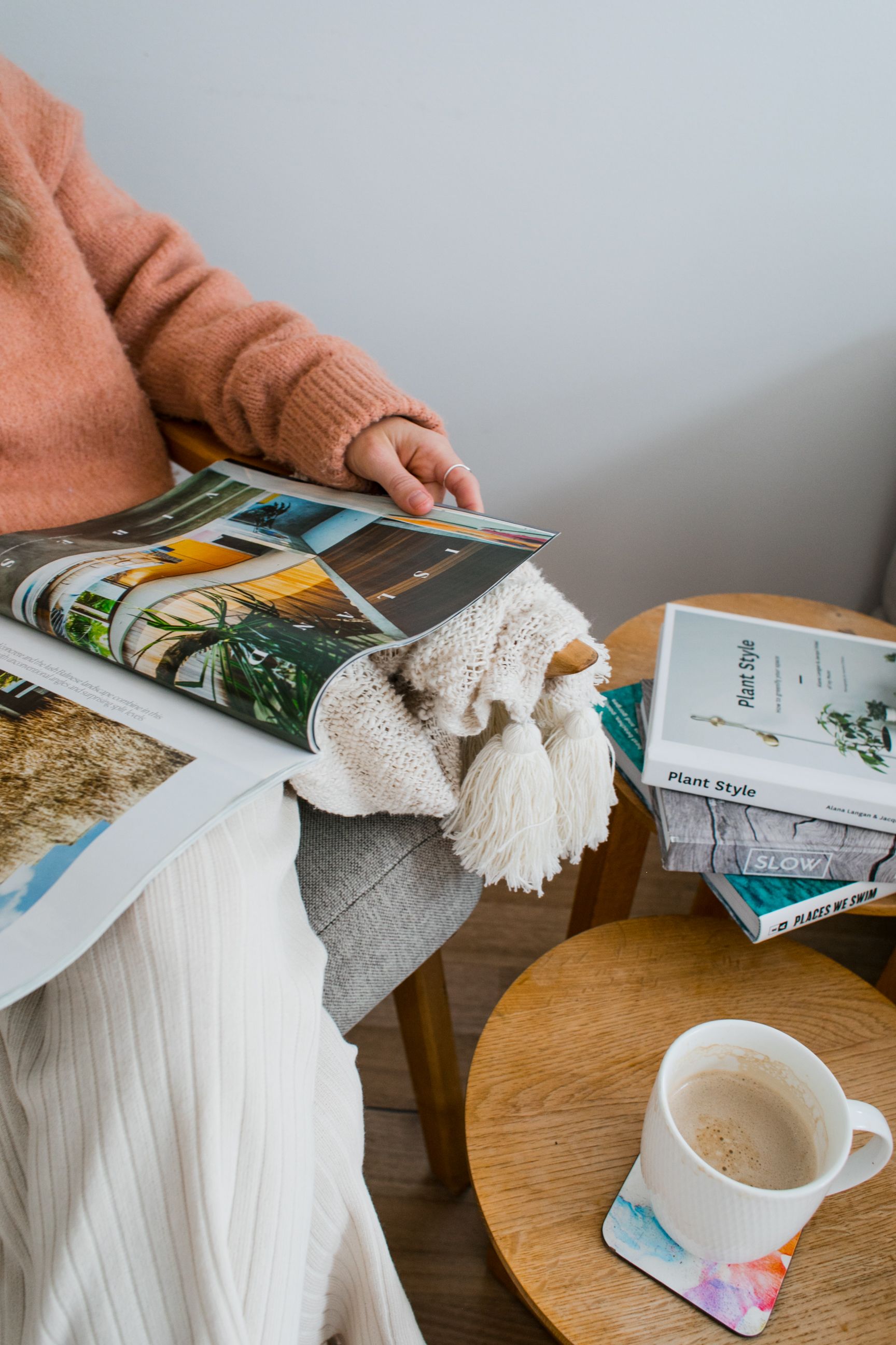 Una mujer está sentada en una silla leyendo una revista junto a una taza de café.