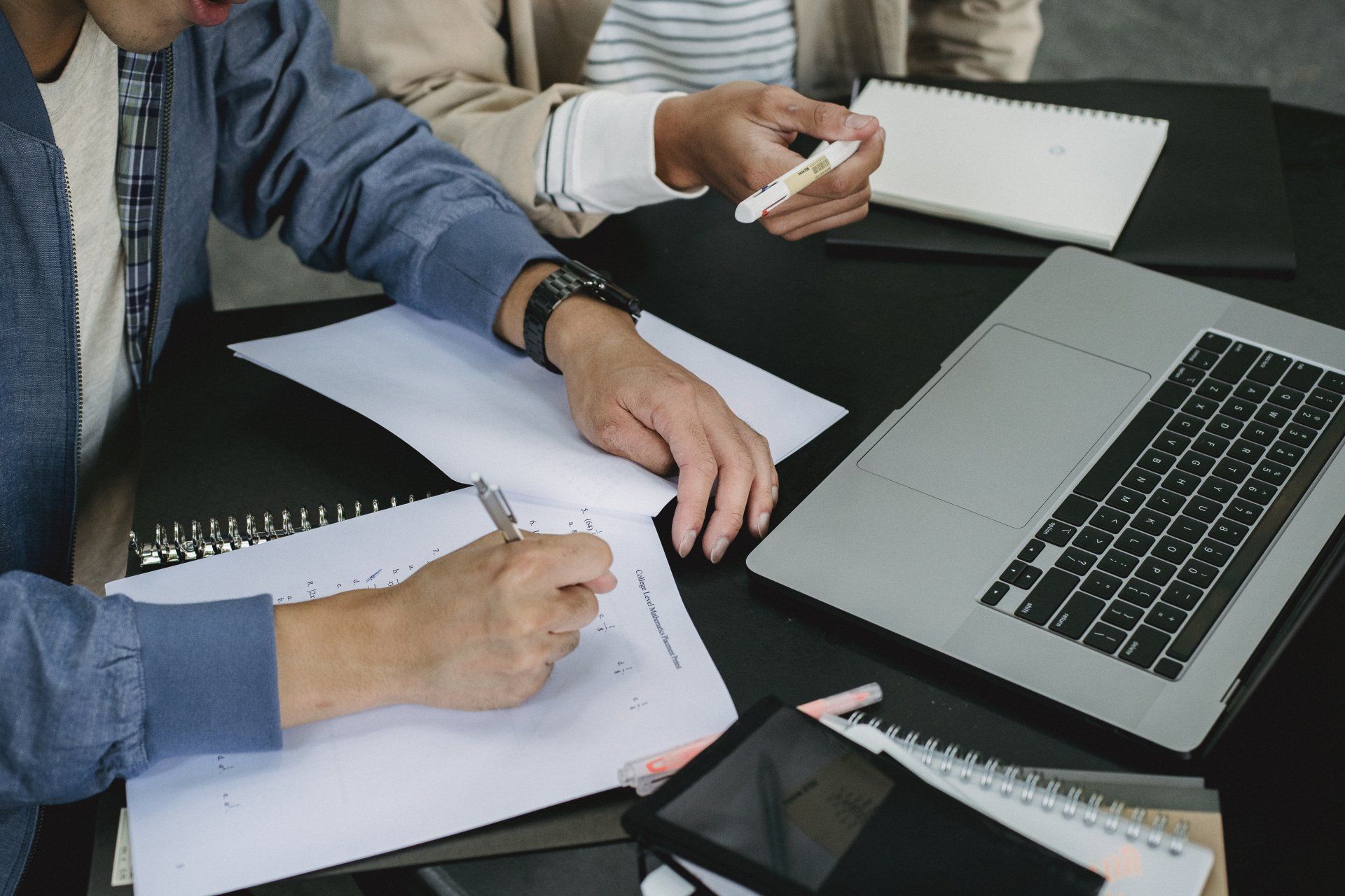 Two people collaborate at a table with a laptop, notebooks, and pens, focusing on writing and working on documents.
