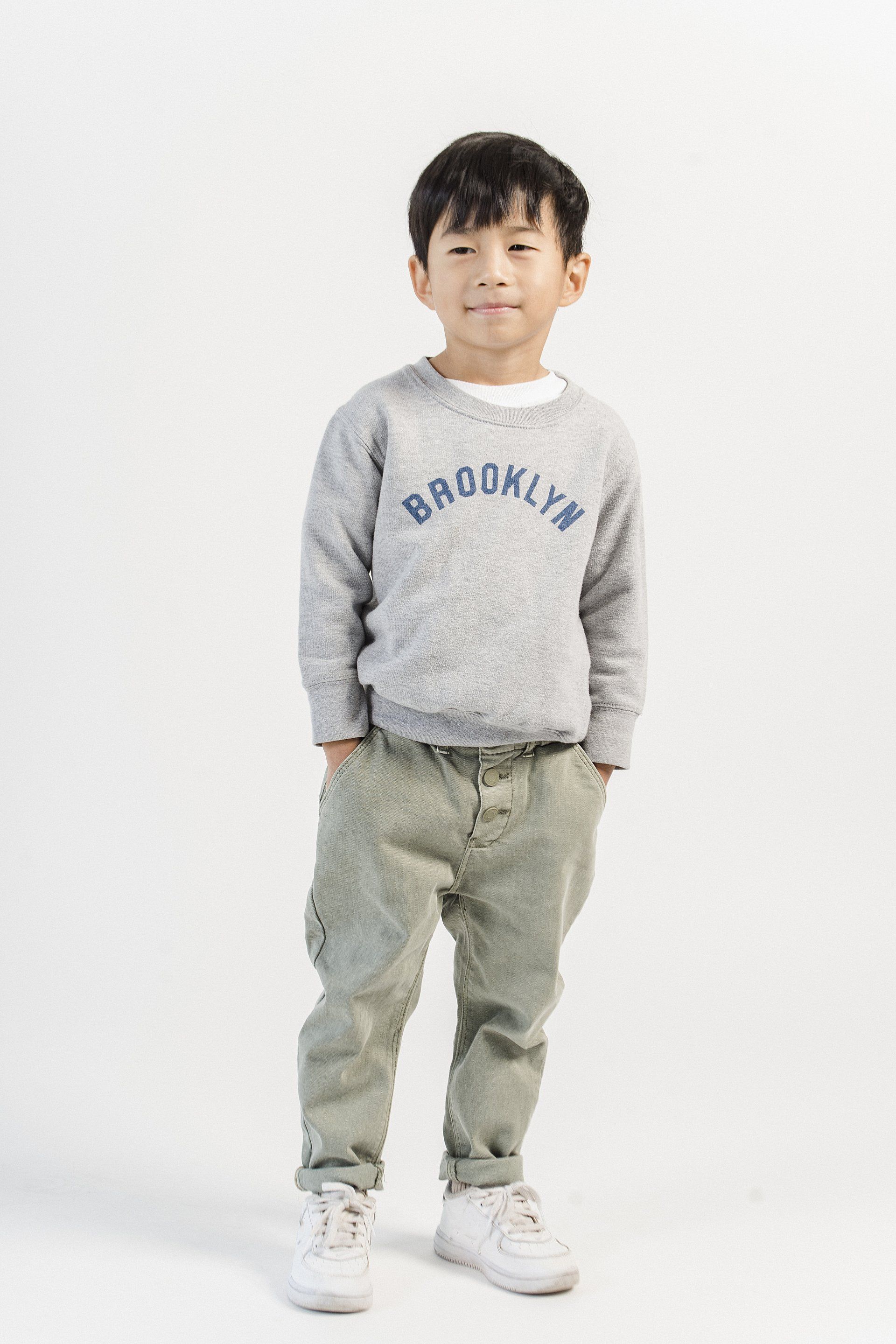 A young boy wearing a gray sweatshirt and khaki pants is standing with his hands in his pockets.