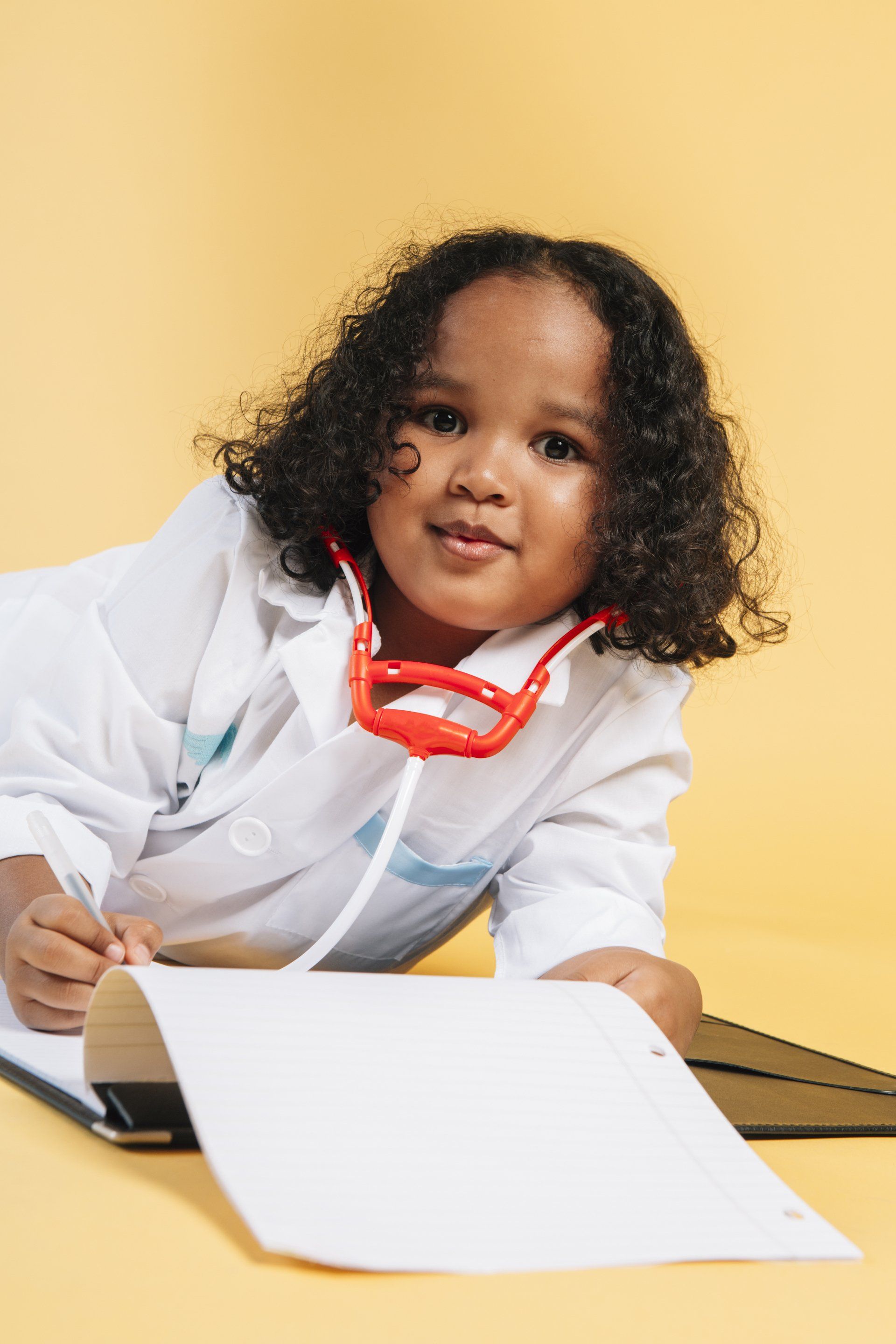 A little girl dressed as a doctor is laying on the floor with a stethoscope around her neck.