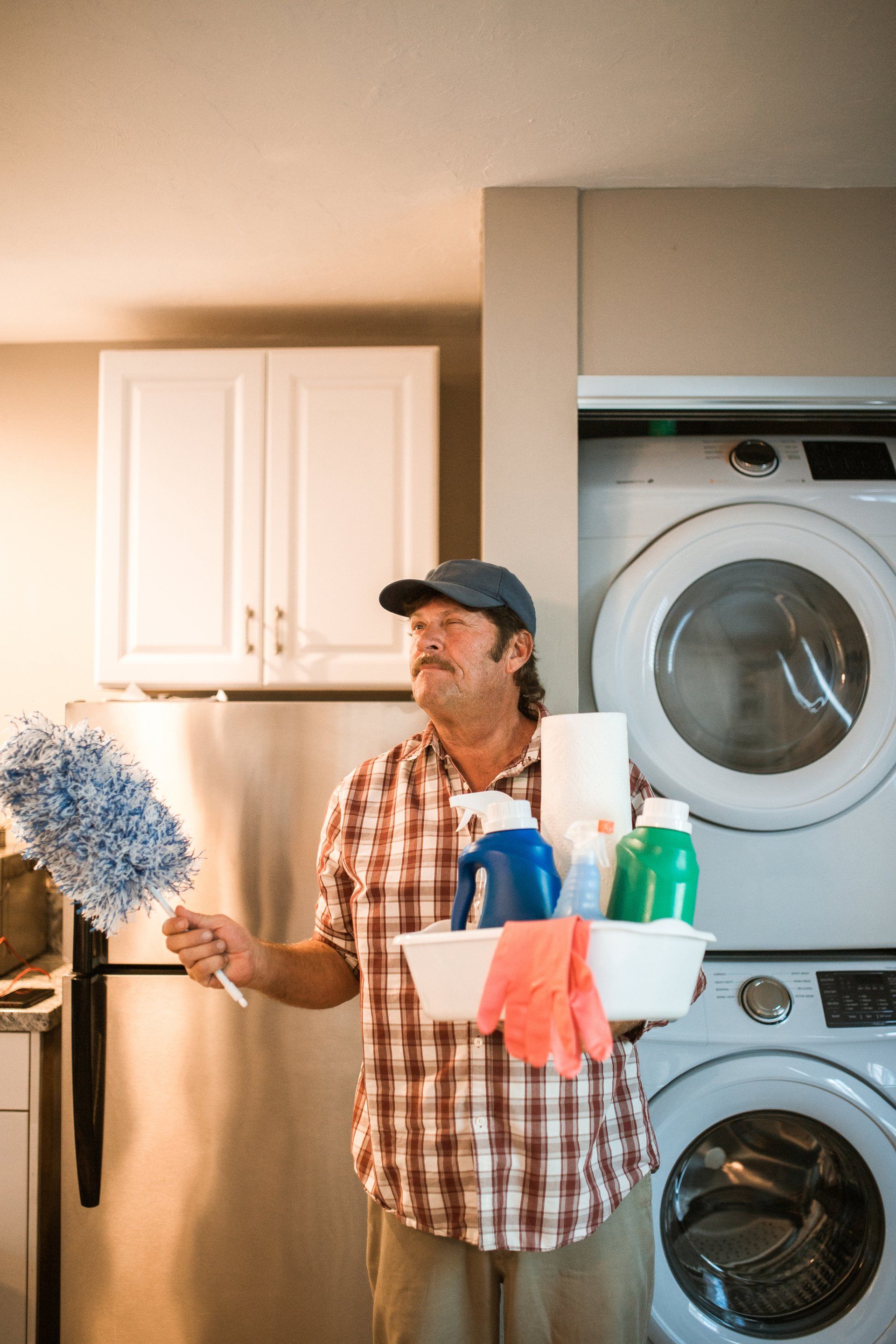 A man is standing in a laundry room holding a duster and a basket of cleaning supplies.