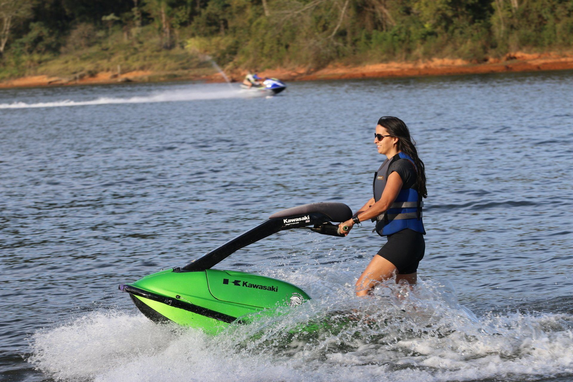 Woman on a green jet ski on a lake, wearing a life vest and sunglasses. Another jet ski in the background.